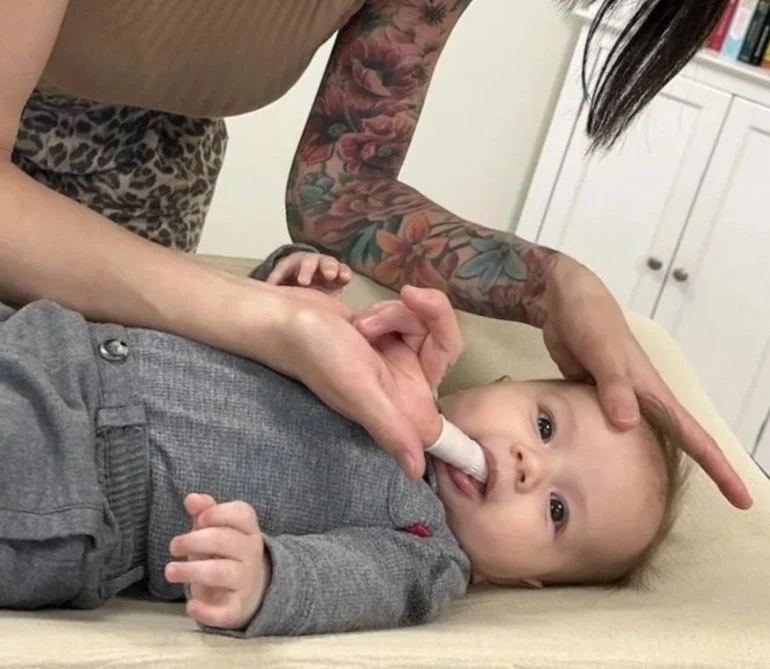 A baby with a pacifier in its mouth lying on a changing table, being gently held by a woman with a tattooed arm. The woman’s other hand is placed on the baby's head, and she is wearing a leopard print top. The background shows a white cabinet and books.