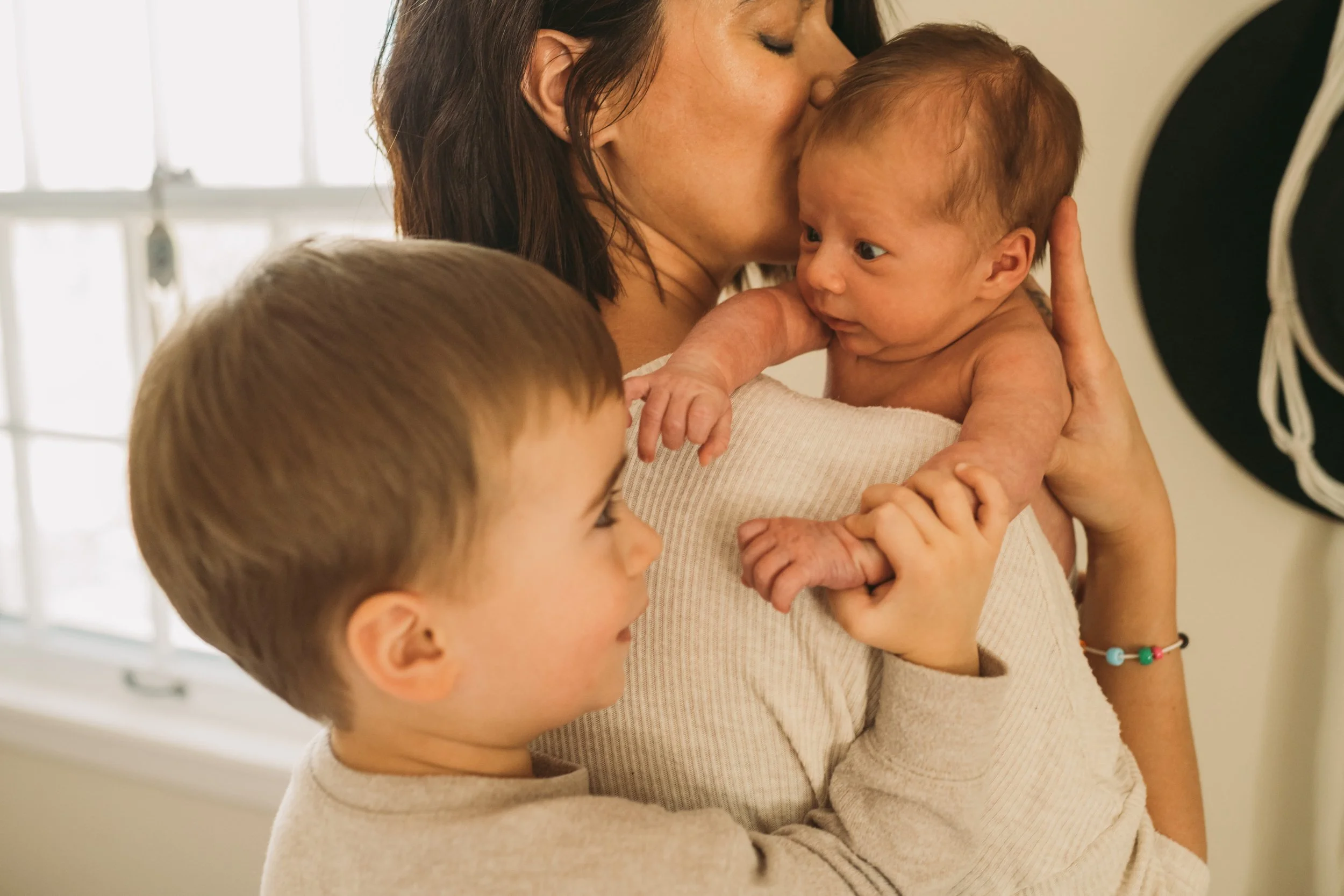 A woman holding a baby close while kissing the baby's forehead. Two other children are nearby, one looking at the woman and baby, the other facing away. Indoor setting with natural light.