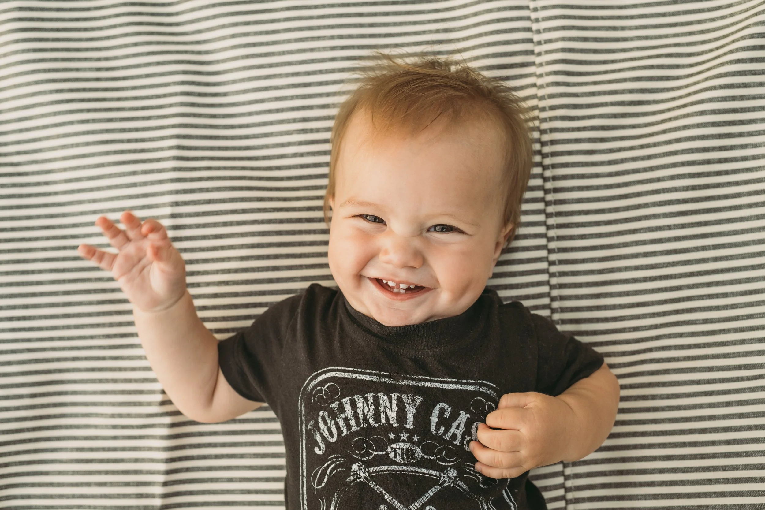 A young boy with light brown hair smiling and waving, wearing a black T-shirt with a graphic design, lying on a black and white striped fabric background.