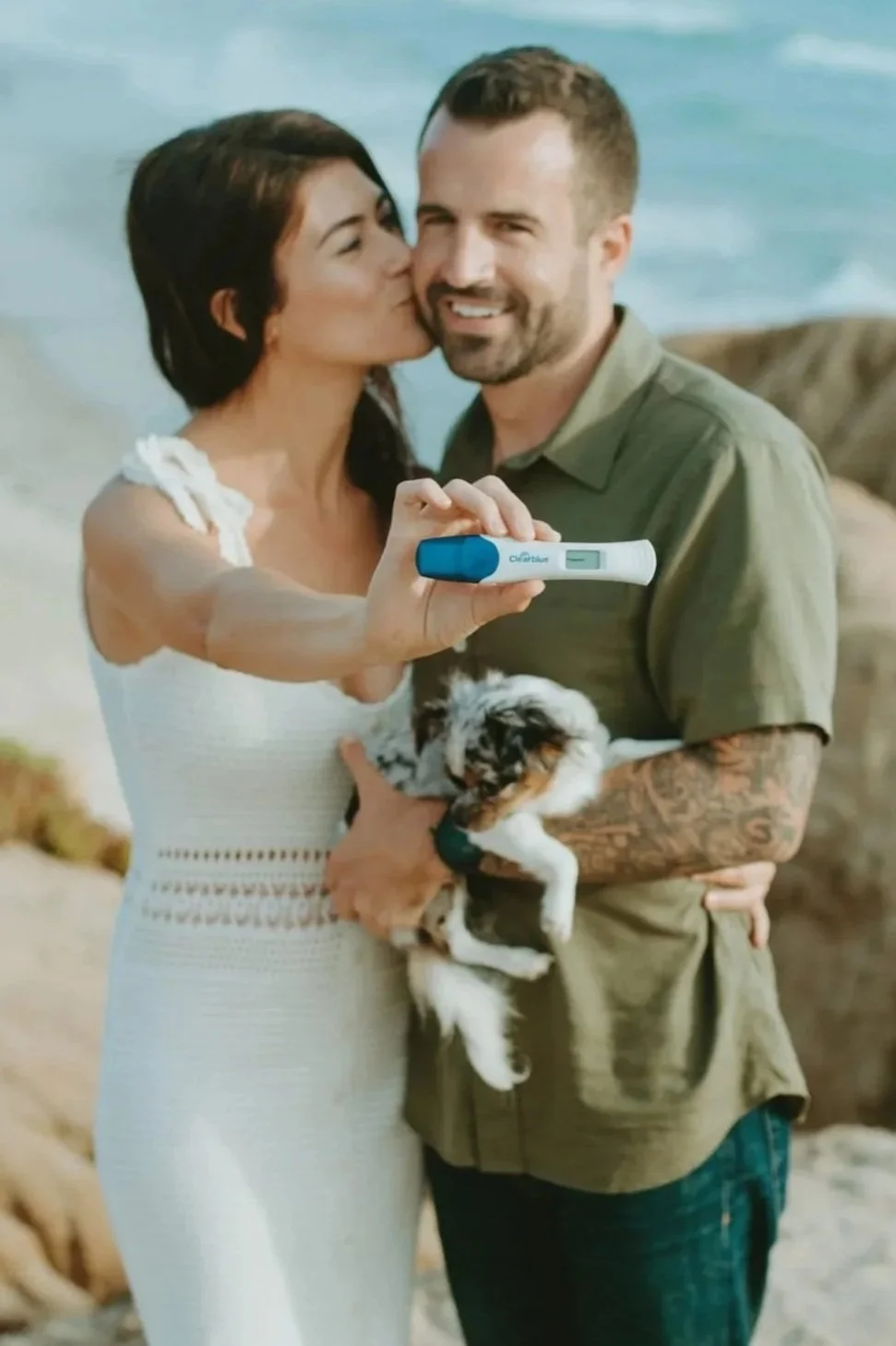 A couple stands on a beach, taking a selfie with a pregnancy test. The woman holds a small puppy, and the man has tattoos on his arm. The background shows sand and water, indicating a seaside setting.