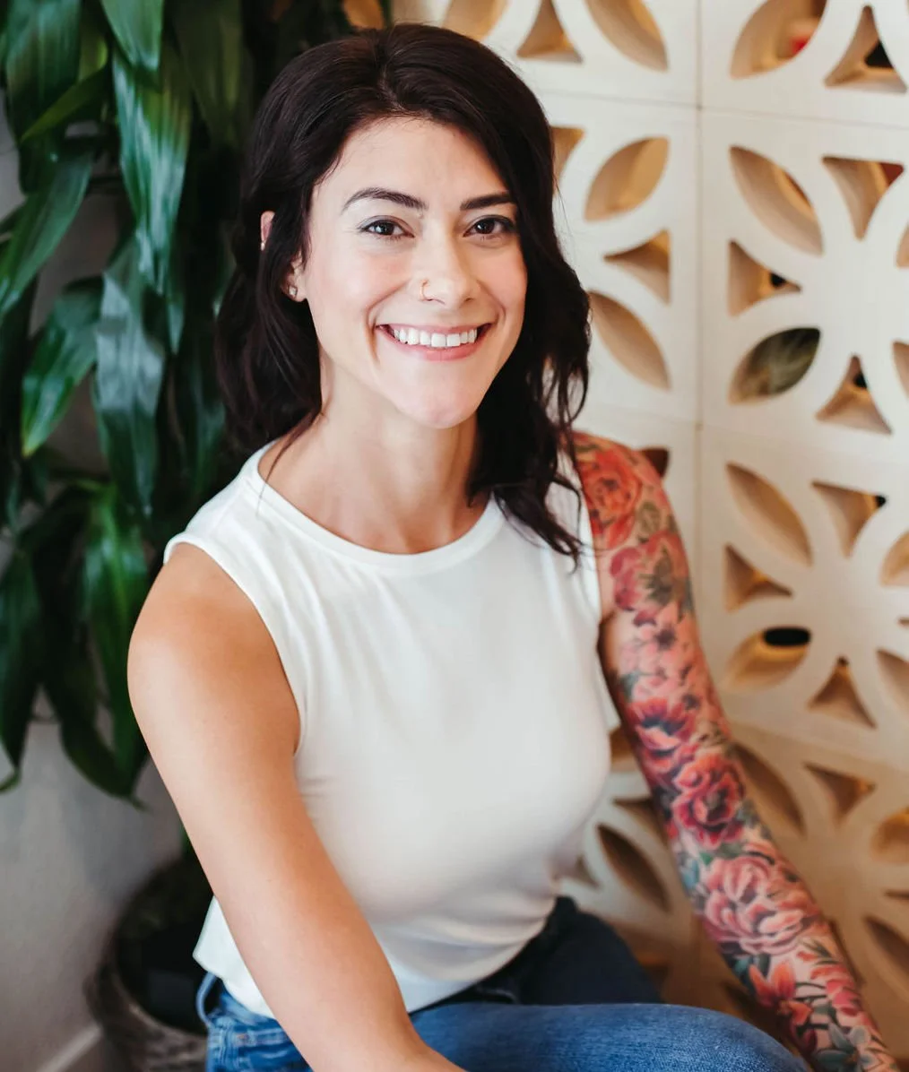 A woman with dark hair and tattoos on her right arm, smiling and sitting indoors with a decorative wooden wall and green plant in the background.