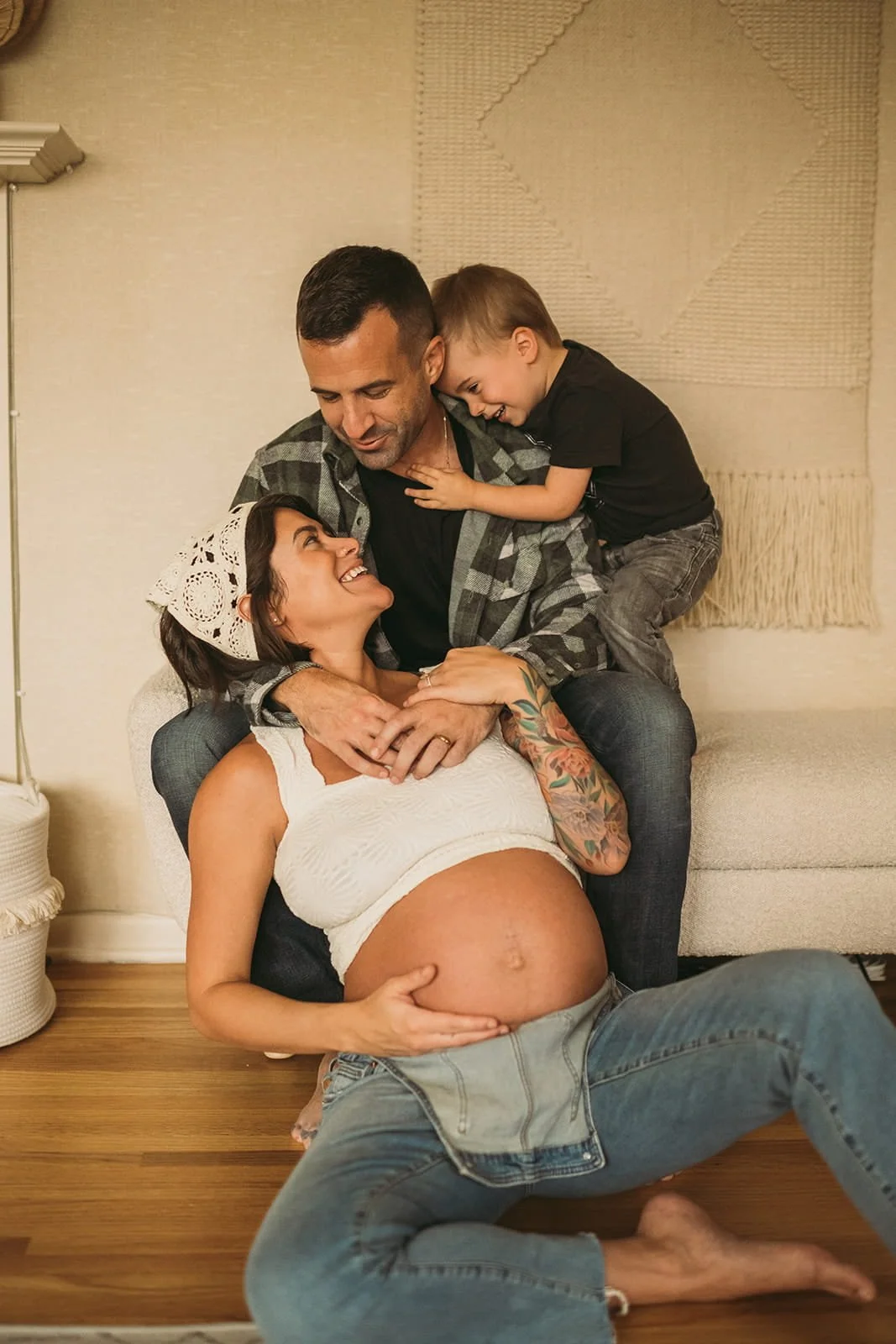 A pregnant woman with tattoos on her arm sitting on the floor smiling up at a man and two children, all gathered around her with happy expressions. The woman holds her pregnant belly, and the man and children are interacting affectionately, creating a warm family moment.