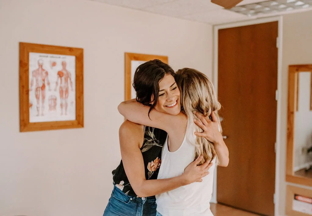 Two women hugging and smiling indoors, with medical posters on the wall behind them.