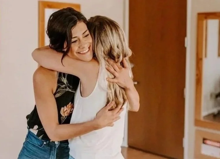 Two women hugging warmly, smiling, in a cozy room.