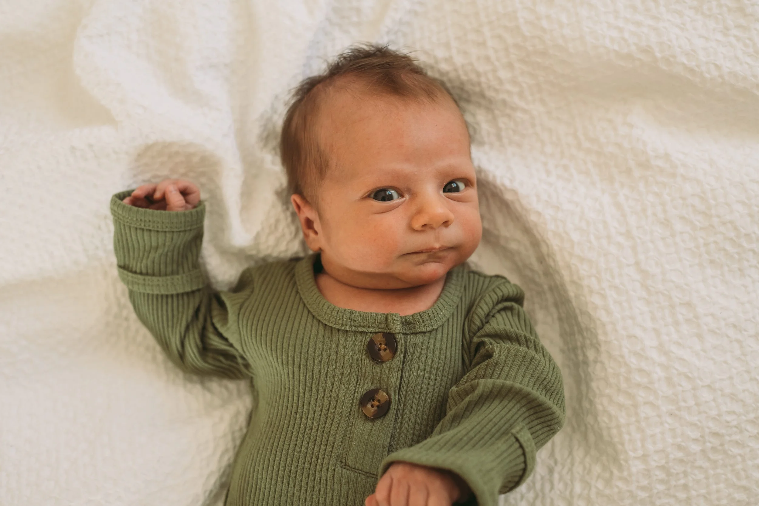 Close-up of a baby lying on a soft, cream-colored blanket, wearing a green ribbed outfit with large brown buttons, looking directly at the camera.