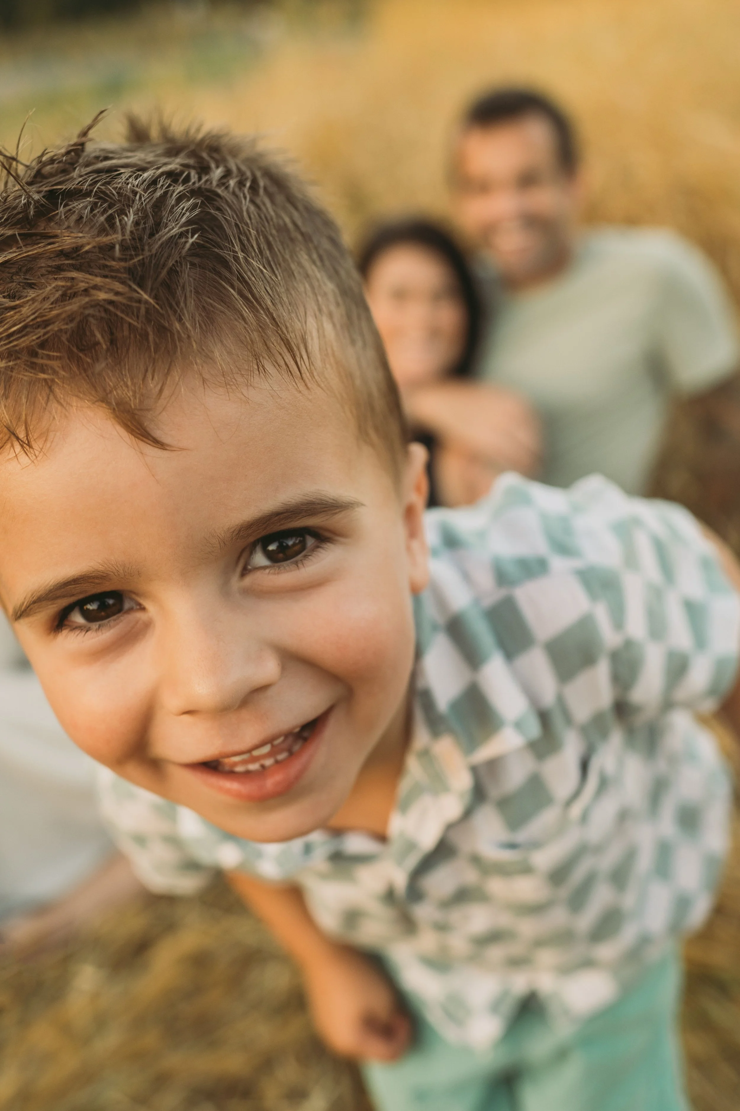 Close-up of a smiling young boy with brown hair and brown eyes, wearing a light green and white checkered shirt, outdoors with blurred background.