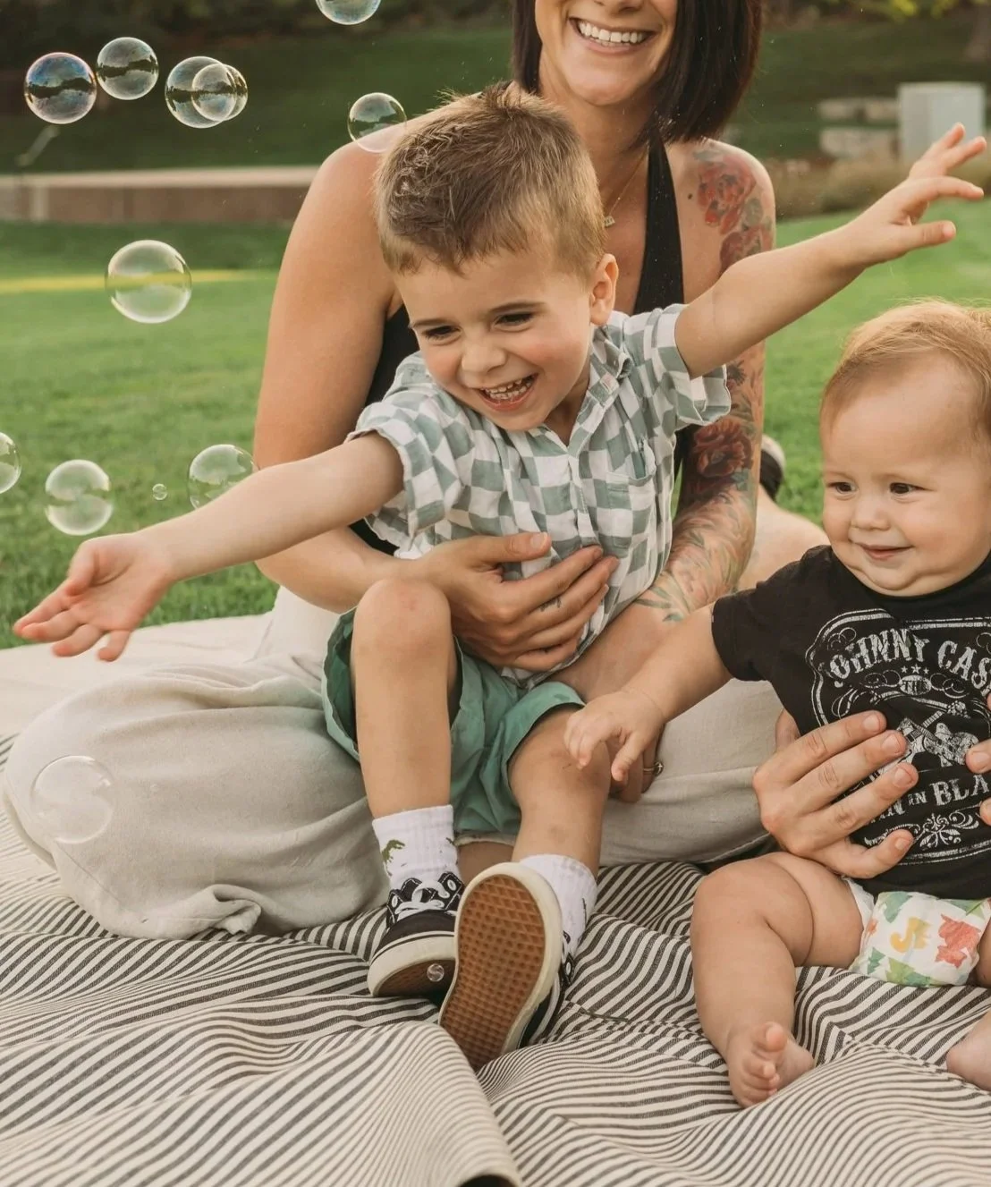 A woman with tattoos on her arm and shoulder sitting on a blanket in a park, playing with two young children, one boy and one girl, while bubbles float around them. All three are smiling and enjoying the moment.