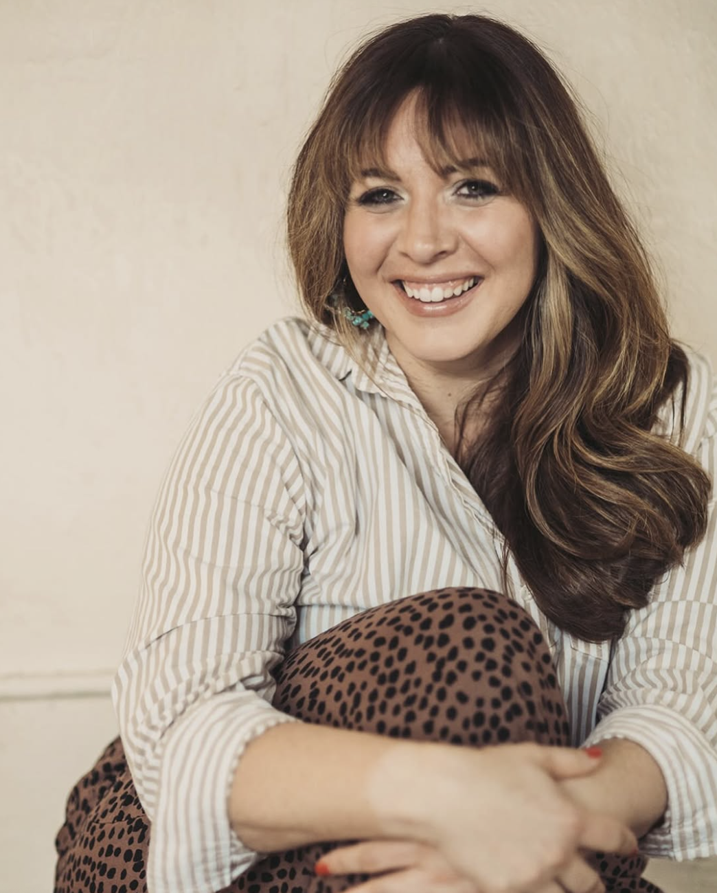 A smiling woman with long brown hair, wearing a beige and white striped shirt and leopard-print pants, sitting with her arms wrapped around her knees against a plain beige wall.