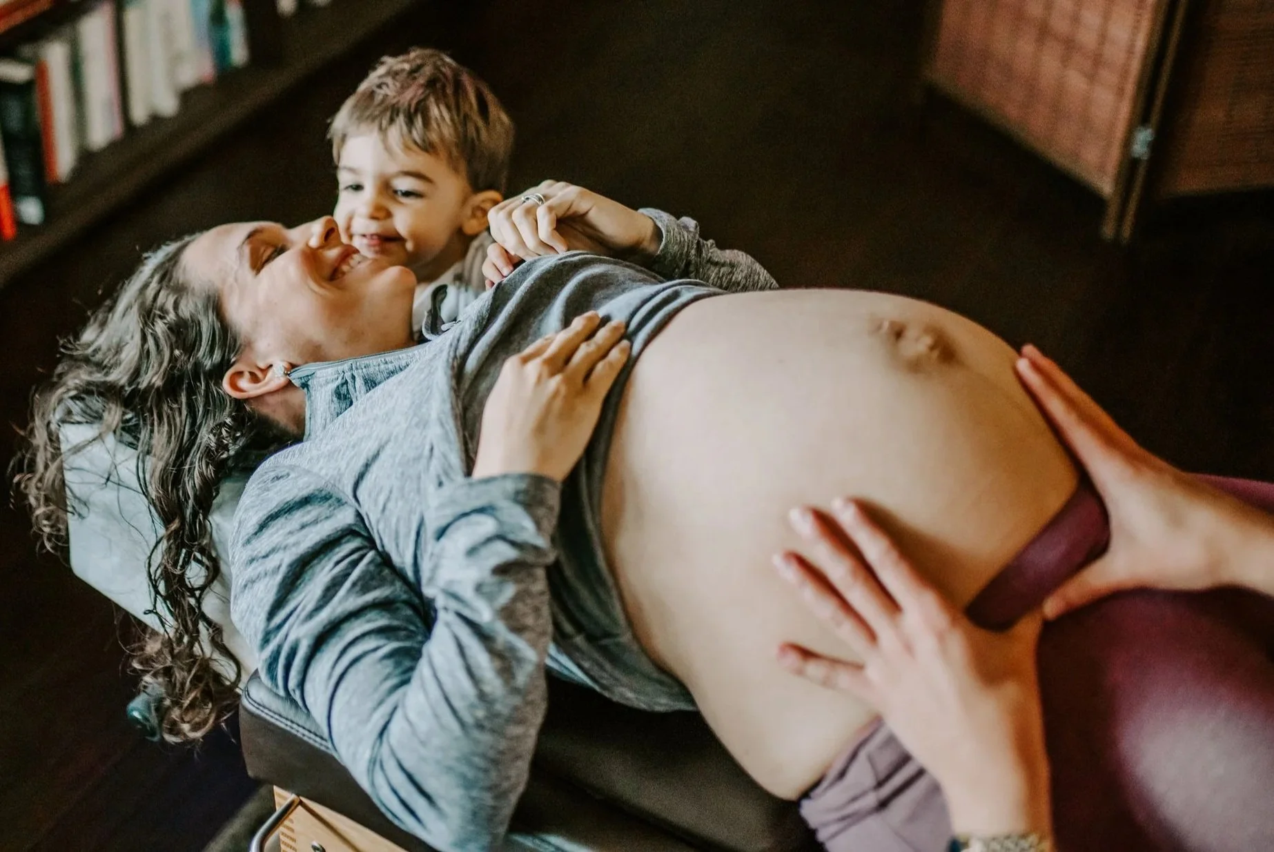 A pregnant woman lying on a bed with a young boy, both smiling and holding hands.