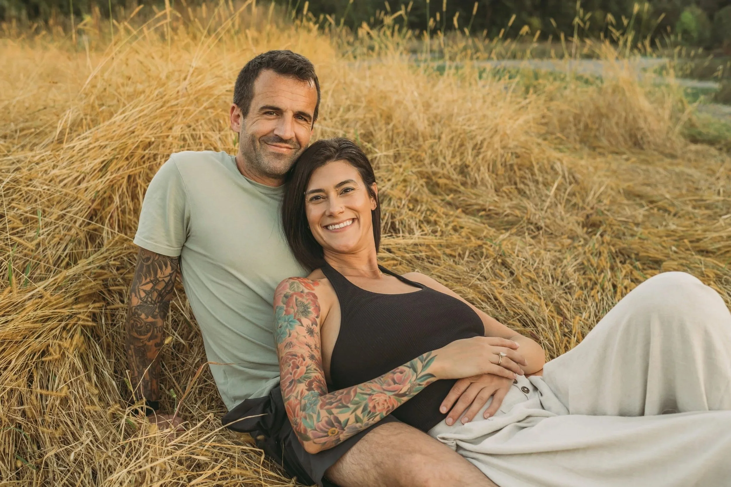 A smiling man and woman sitting in a field of tall golden grass, with the woman resting her hand on her stomach, possibly indicating pregnancy, and both enjoying a relaxed moment outdoors.