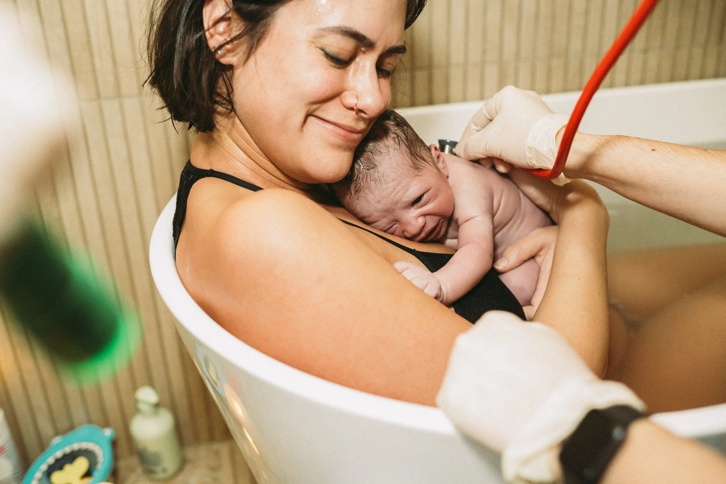 A woman holding a newborn baby during a home birth, with medical gloves and a medical professional taking care of the baby.