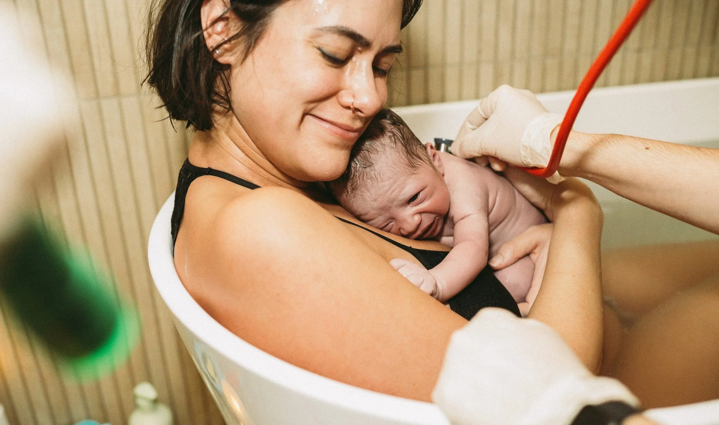 A woman holding a newborn baby in a hospital room, with a healthcare professional attending to the baby.