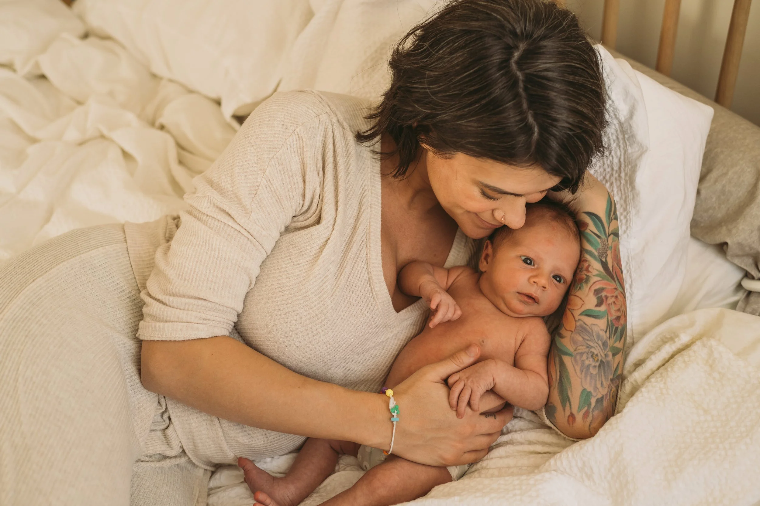 A woman with short dark hair and tattoos on her arm, lying on a bed with white sheets, holding a smiling baby with dark hair and bright eyes.