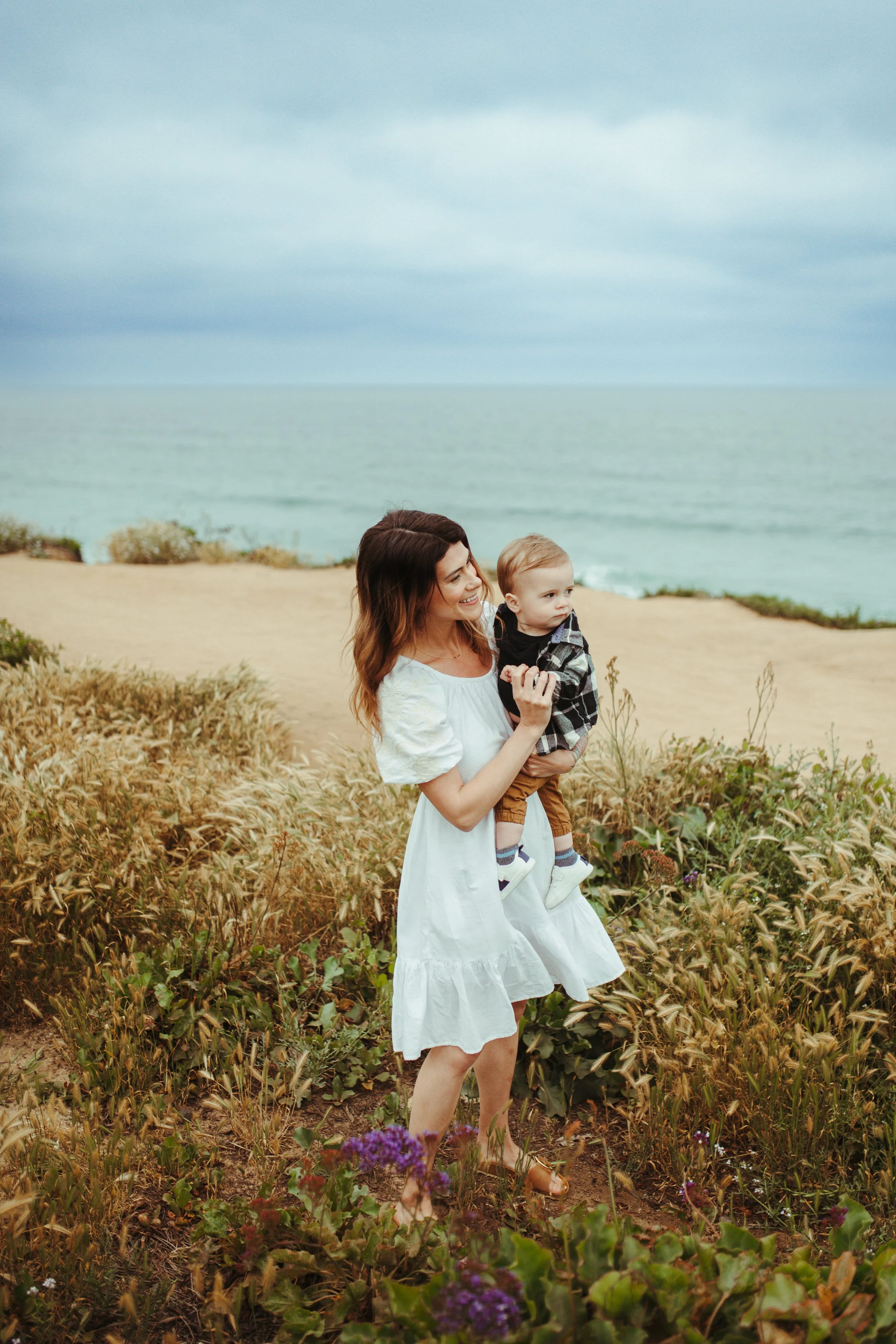 A woman in a white dress holding a young boy in a plaid shirt and brown pants outdoors near a coastal area with ocean and cloudy sky in the background.