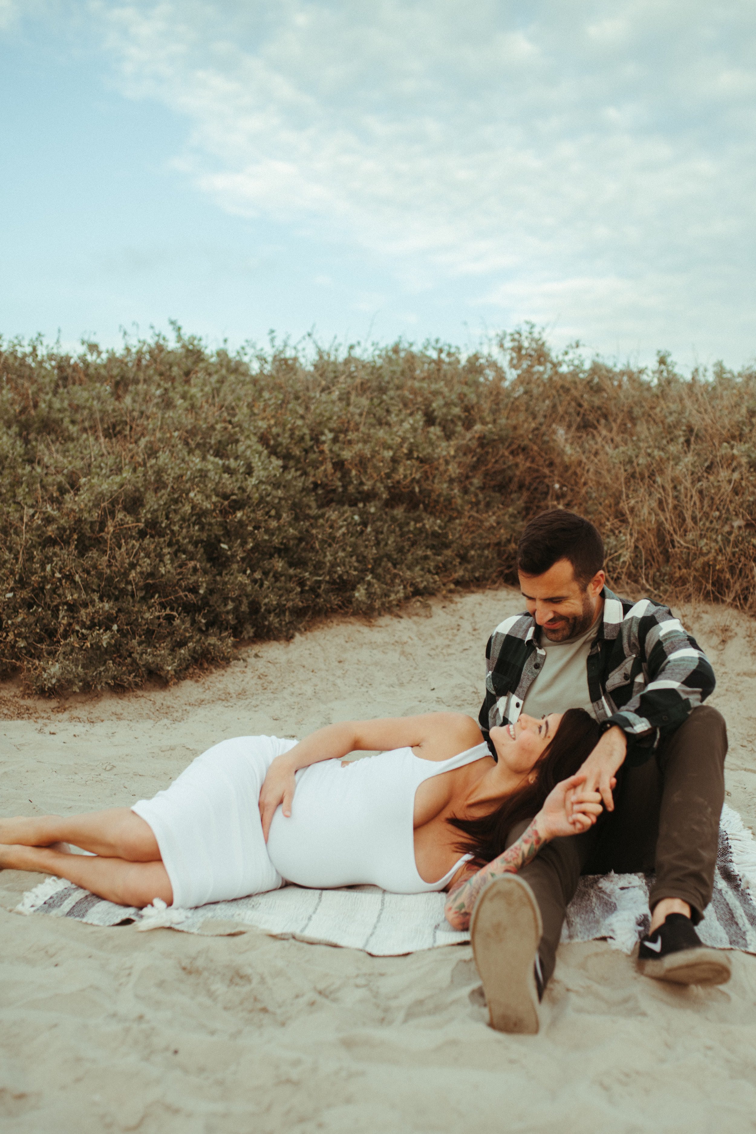 A man and woman lying on a beach, with the man smiling and looking at the woman, who is lying on a towel and smiling back. They are surrounded by sand and bushes, under a partly cloudy sky.