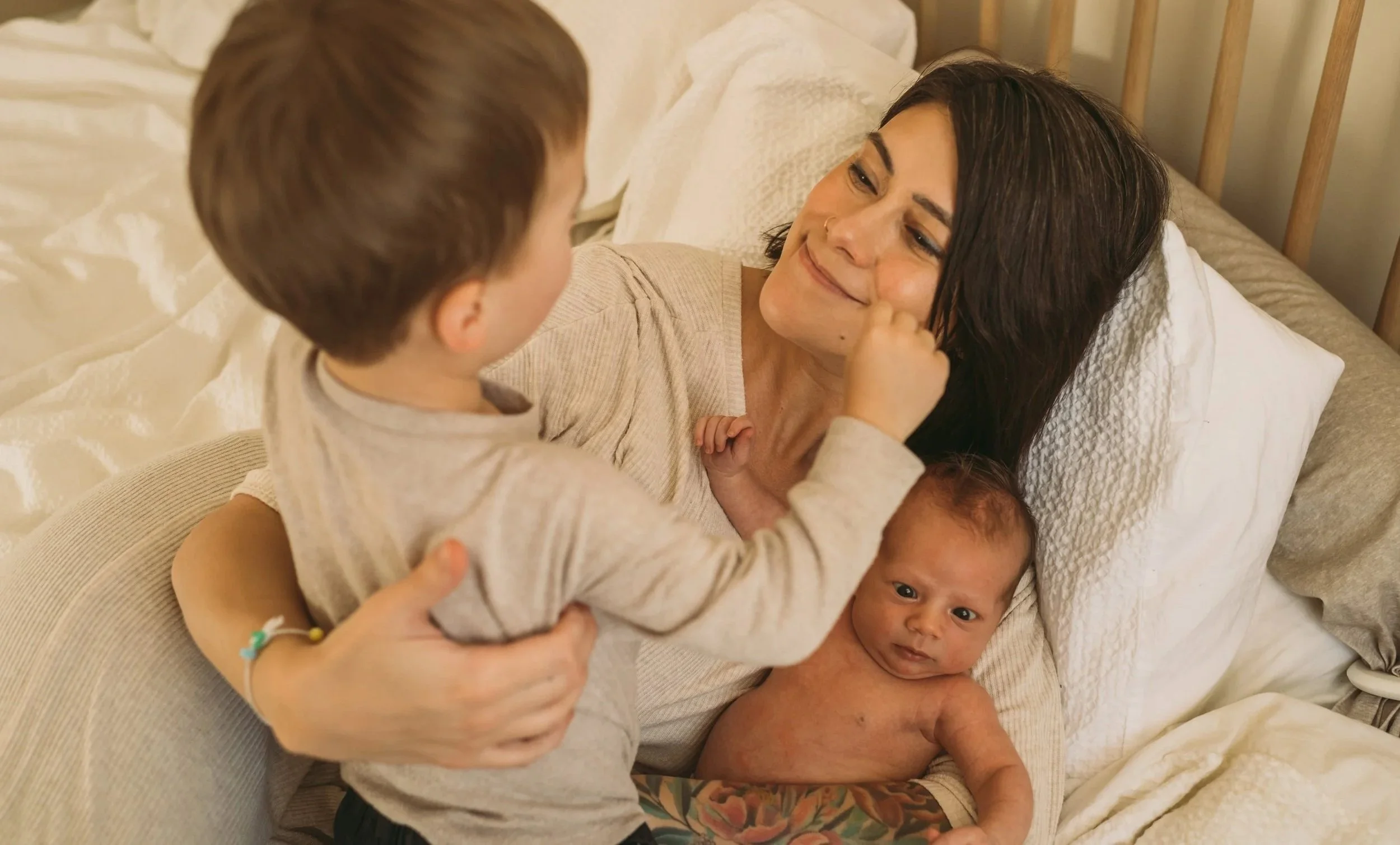 A woman lying on a bed with two young children, one boy sitting on her chest and another baby lying beside her, all smiling and engaging affectionately.