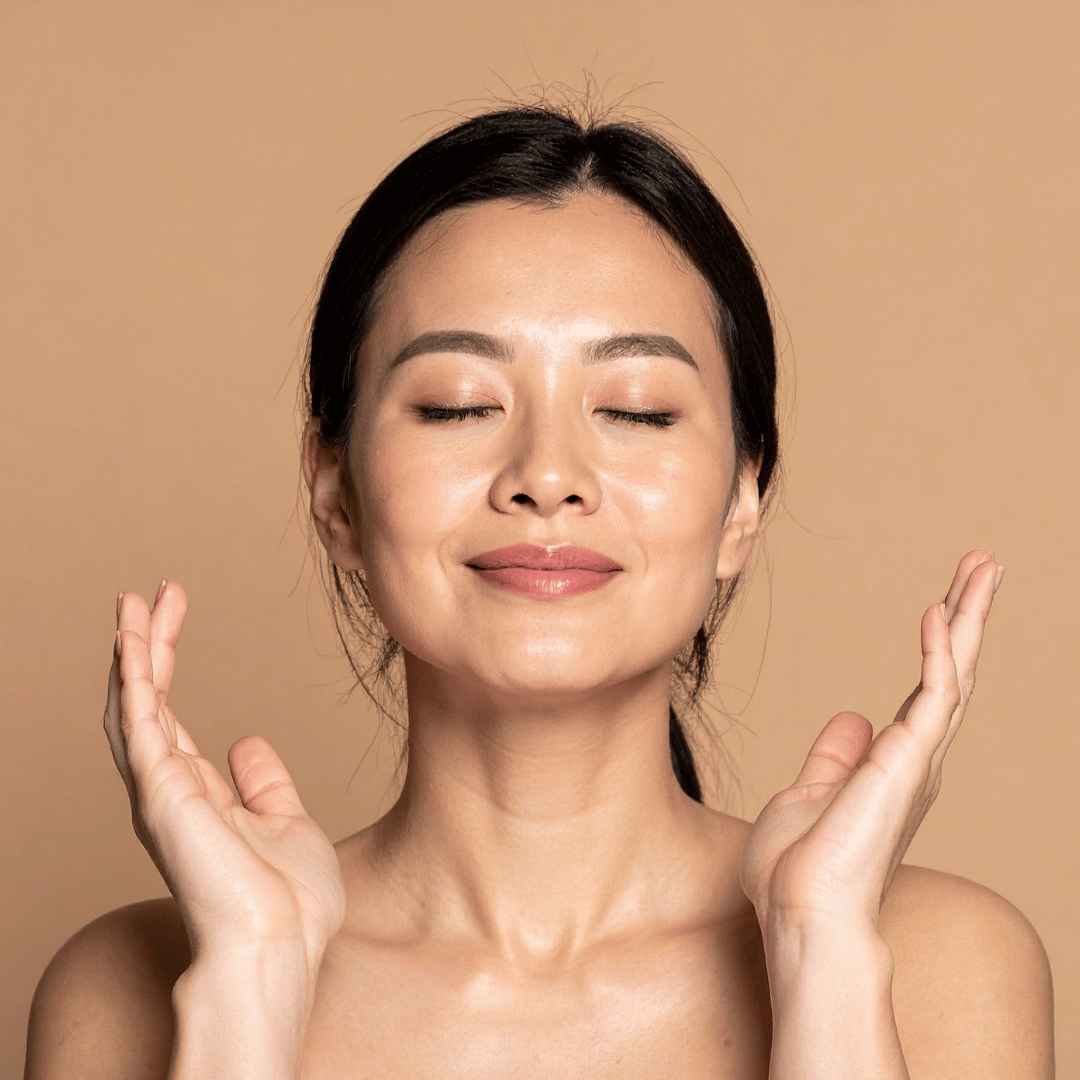 A woman with closed eyes smiling softly, holding her hands near her face in a relaxed pose, against a neutral beige background.