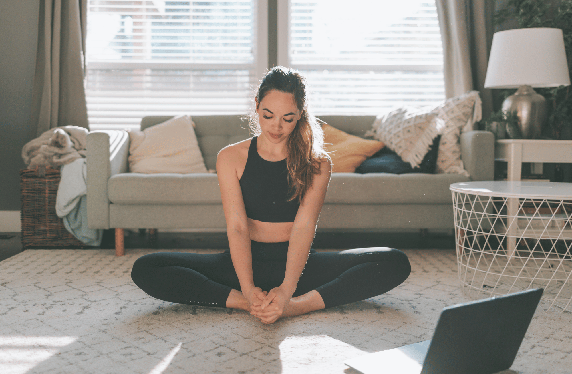 A woman with long brown hair in a ponytail practicing yoga indoors on a beige carpeted floor near a window with blinds. She is sitting cross-legged, wearing a black sports bra and black leggings, with her hands clasped together in front of her. The room has a sofa with pillows and a blanket to her left, and a white side table with decorative objects and a lamp to her right.