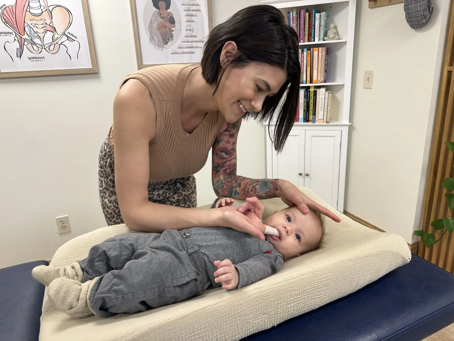 A woman with short dark hair, tattoos on her arm, and a nose piercing, smiles as she gently rubs a baby's head. The baby, lying on a cushioned changing pad, is looking at the camera and appears to be happy. The background features a bookshelf, framed artwork on the wall, and a plant.