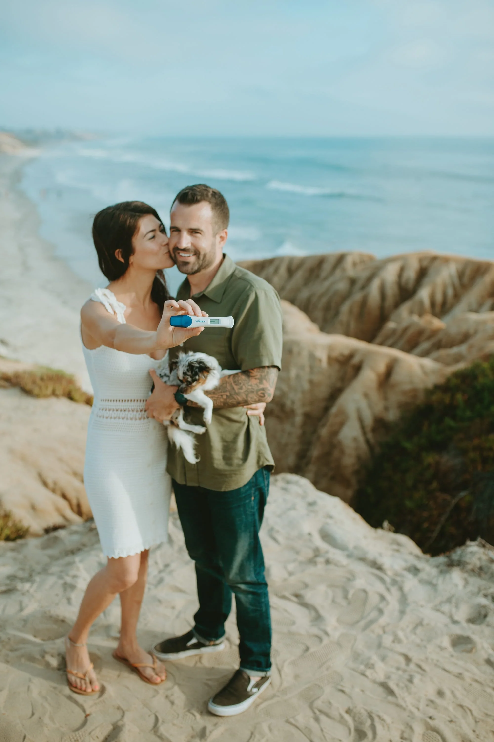 A couple taking a selfie on a beach with a woman kissing a man on the cheek and holding a small dog.