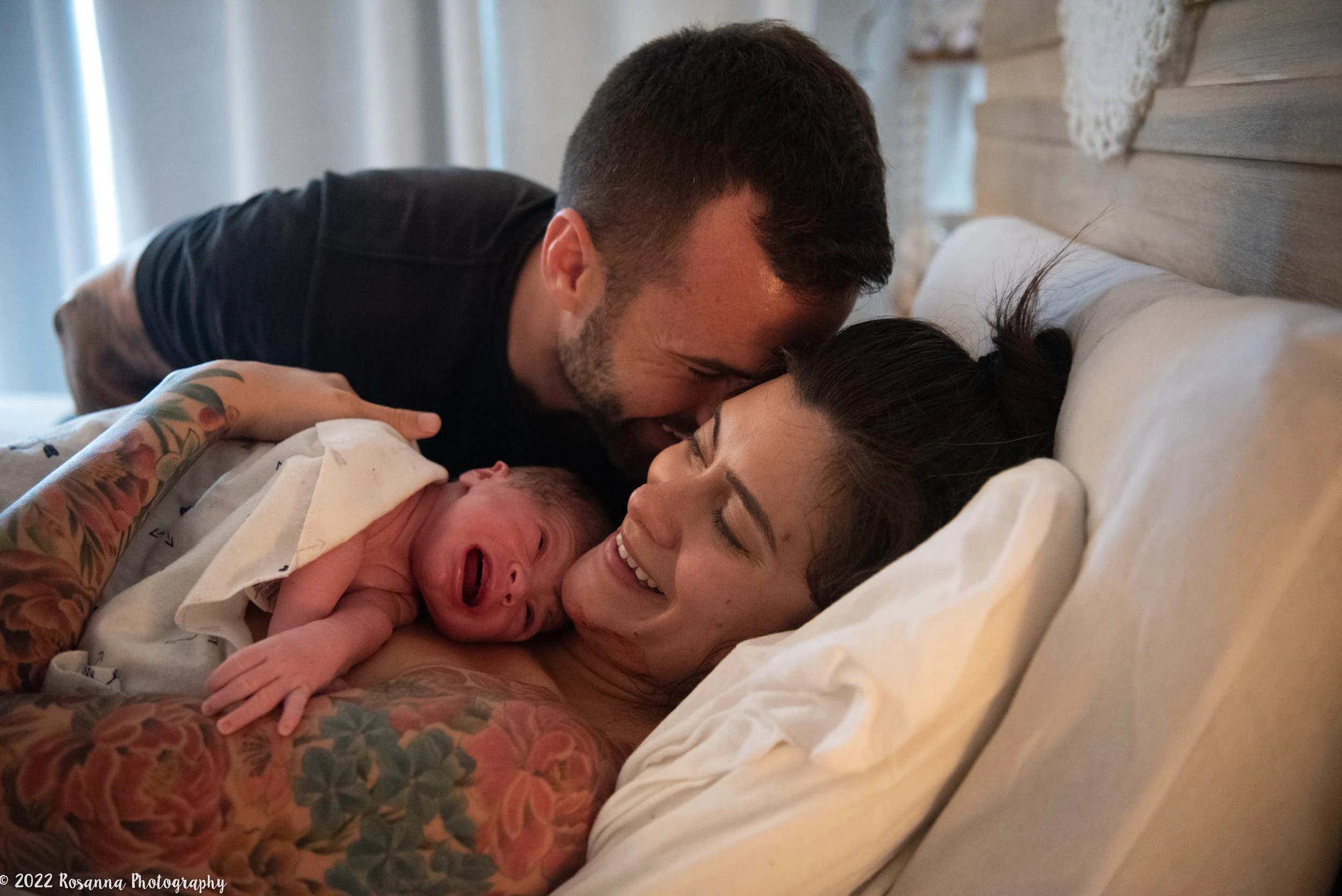 A family in a hospital bed: a woman with tattoos holding a newborn baby, a man leaning over kissing the woman, all smiling and looking happy.