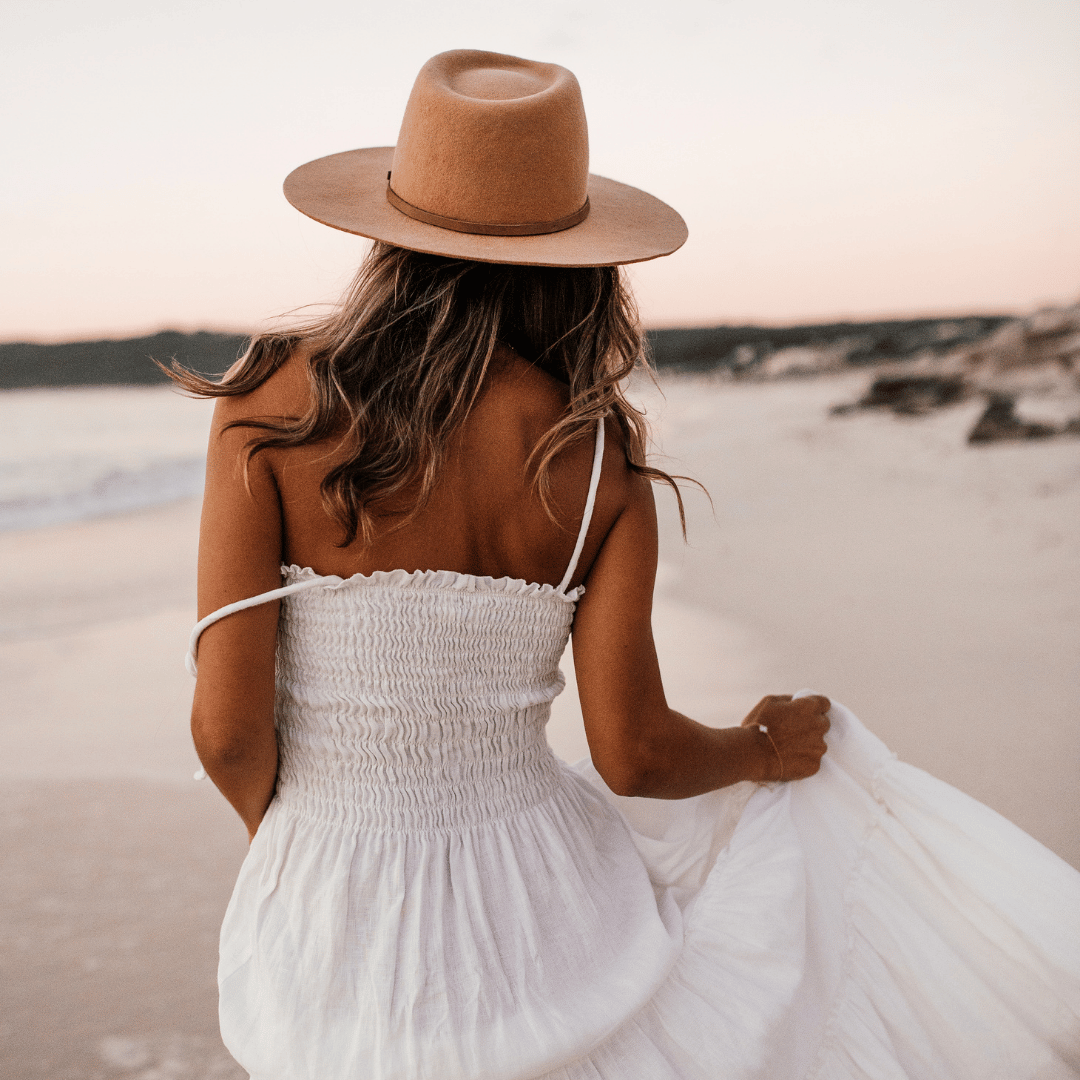 Woman in a white dress and wide-brimmed hat walking along the beach at sunset.