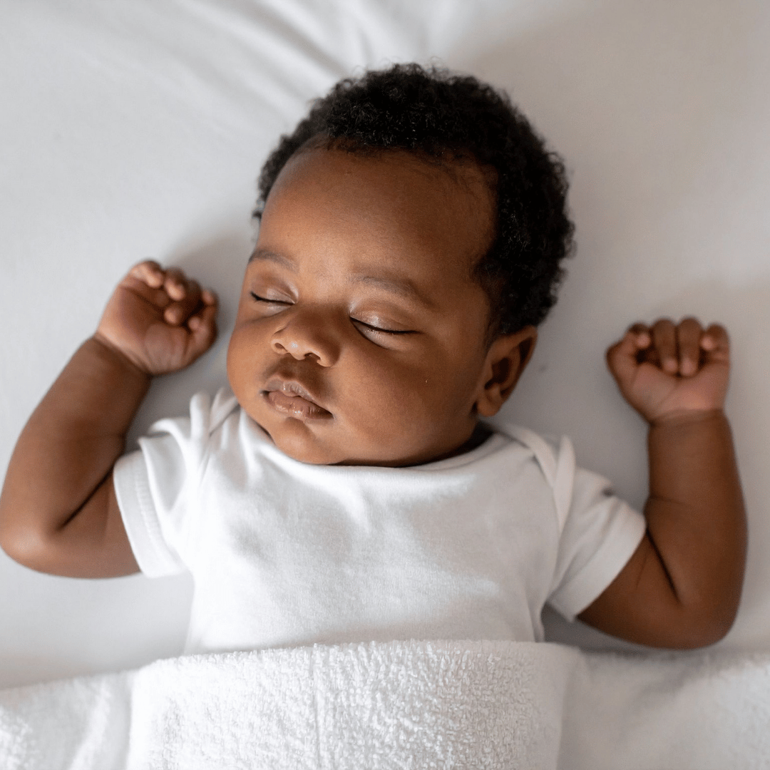 A sleeping infant with dark curly hair lying on a white surface, wearing a white shirt, with eyes closed and arms raised.