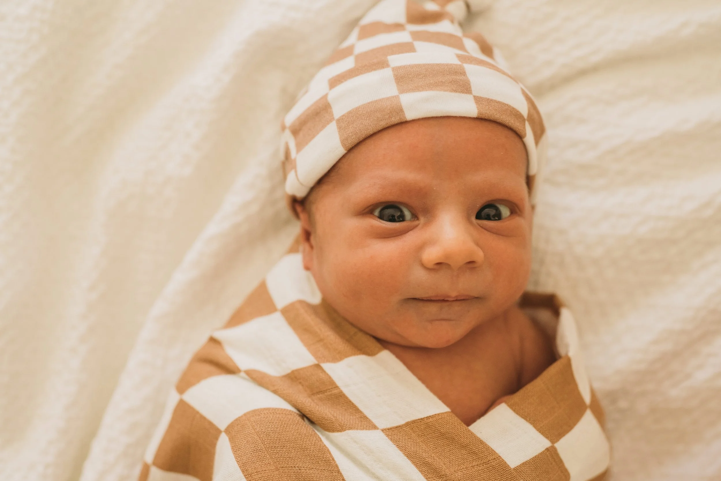Close-up of a baby with a checkered hat and matching outfit, lying on a soft surface, looking at the camera.