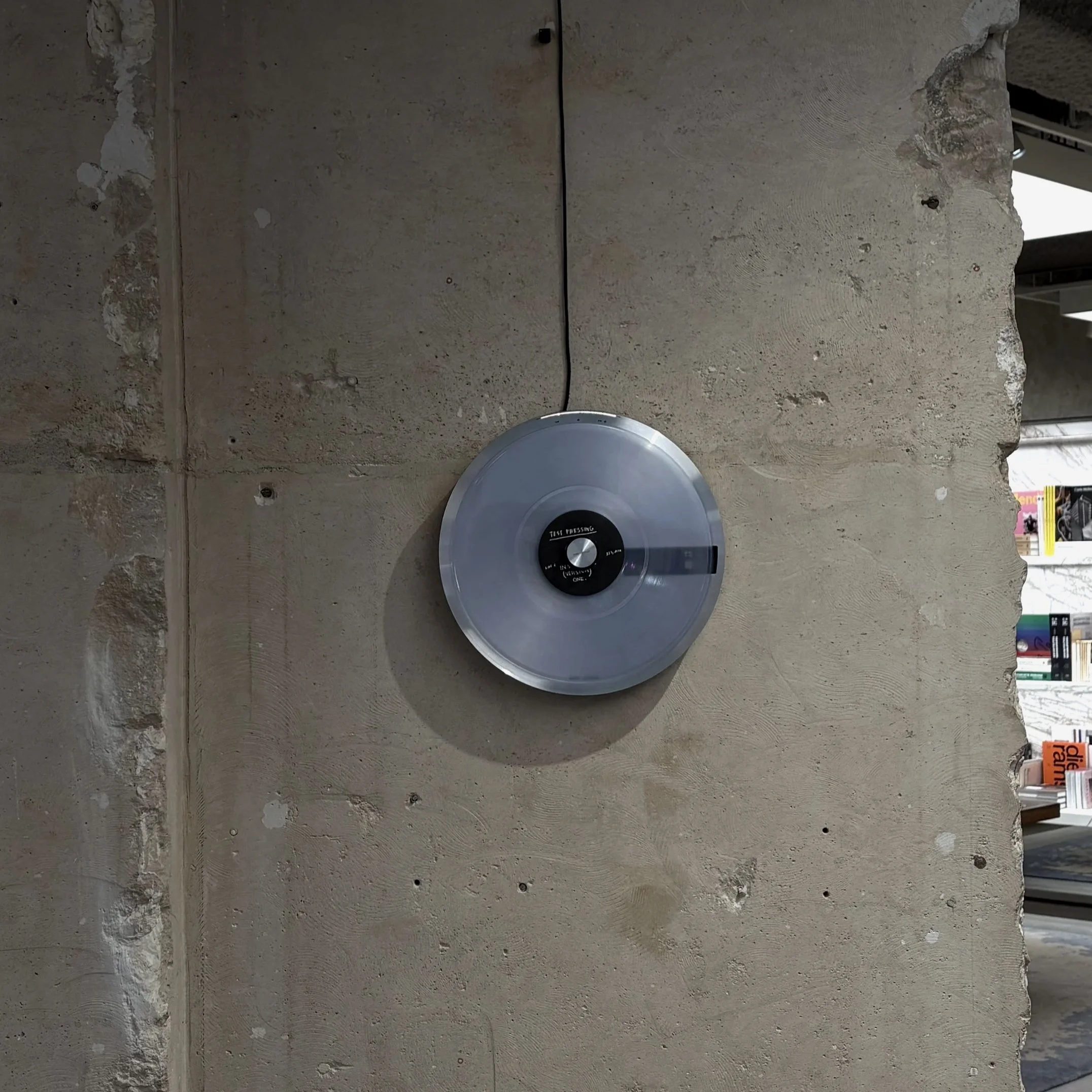 A concrete wall with visible cracks and imperfections, featuring a black cable and a round gray object mounted on it, with store shelves visible in the background.