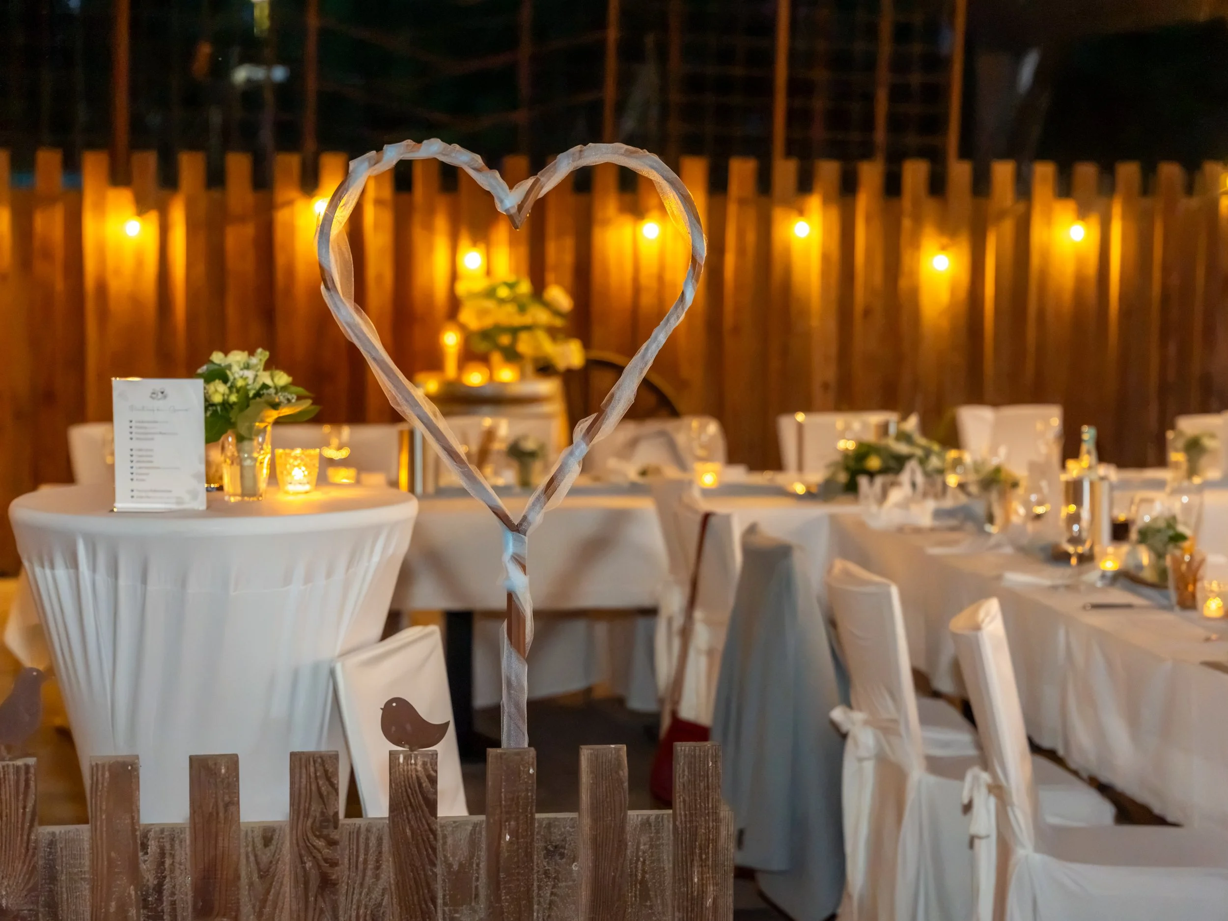 Nighttime wedding reception setup with tables decorated with flowers and candles behind a wooden fence. In the foreground, a heart-shaped decoration made of paper or fabric is attached to a stick, resembling a photo frame.