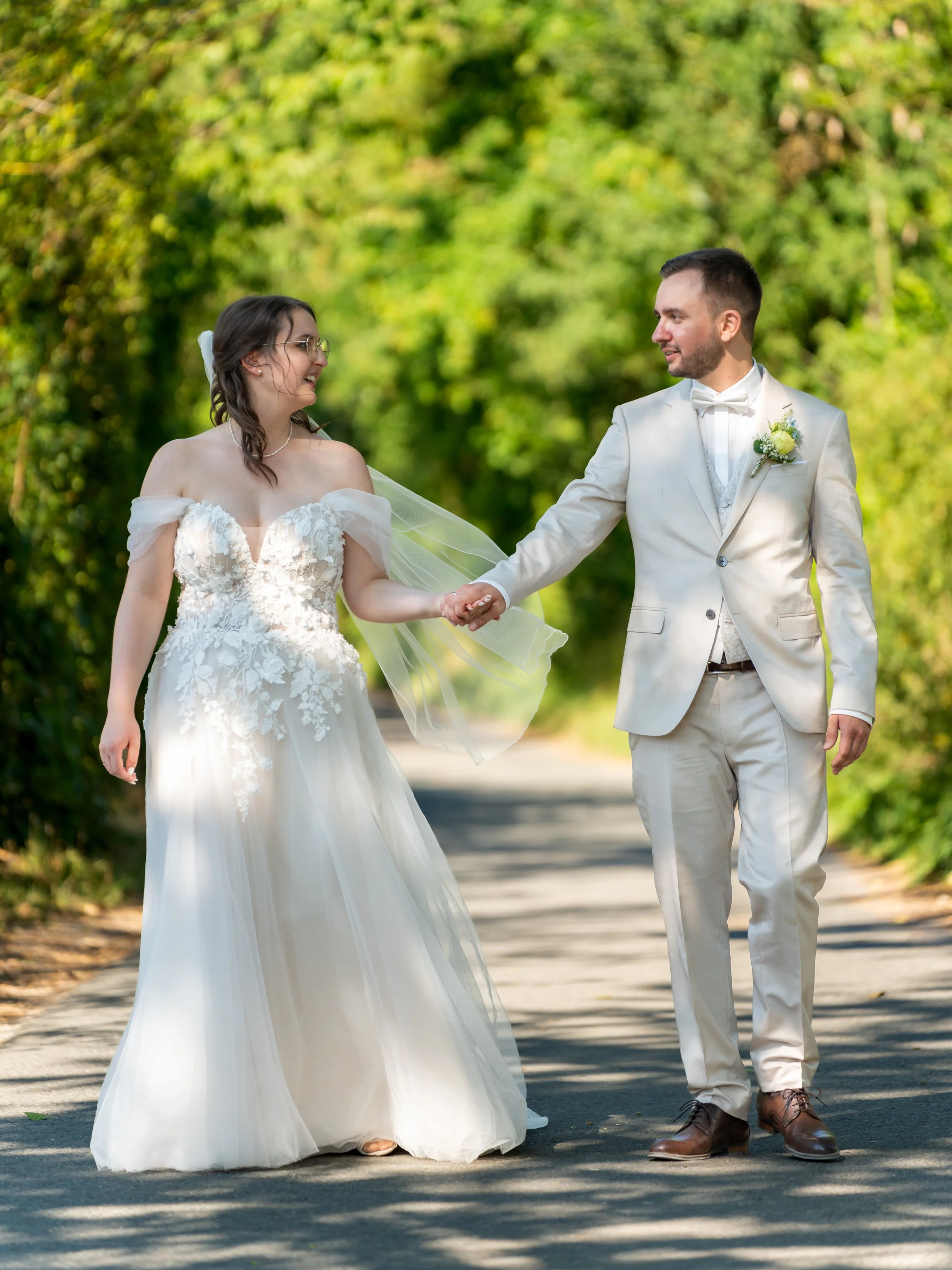 A bride and groom holding hands and walking outdoors on a sunny day, with green trees in the background. The bride is wearing a white wedding dress with lace details and glasses, and the groom is in a light-colored suit with a boutonniere.