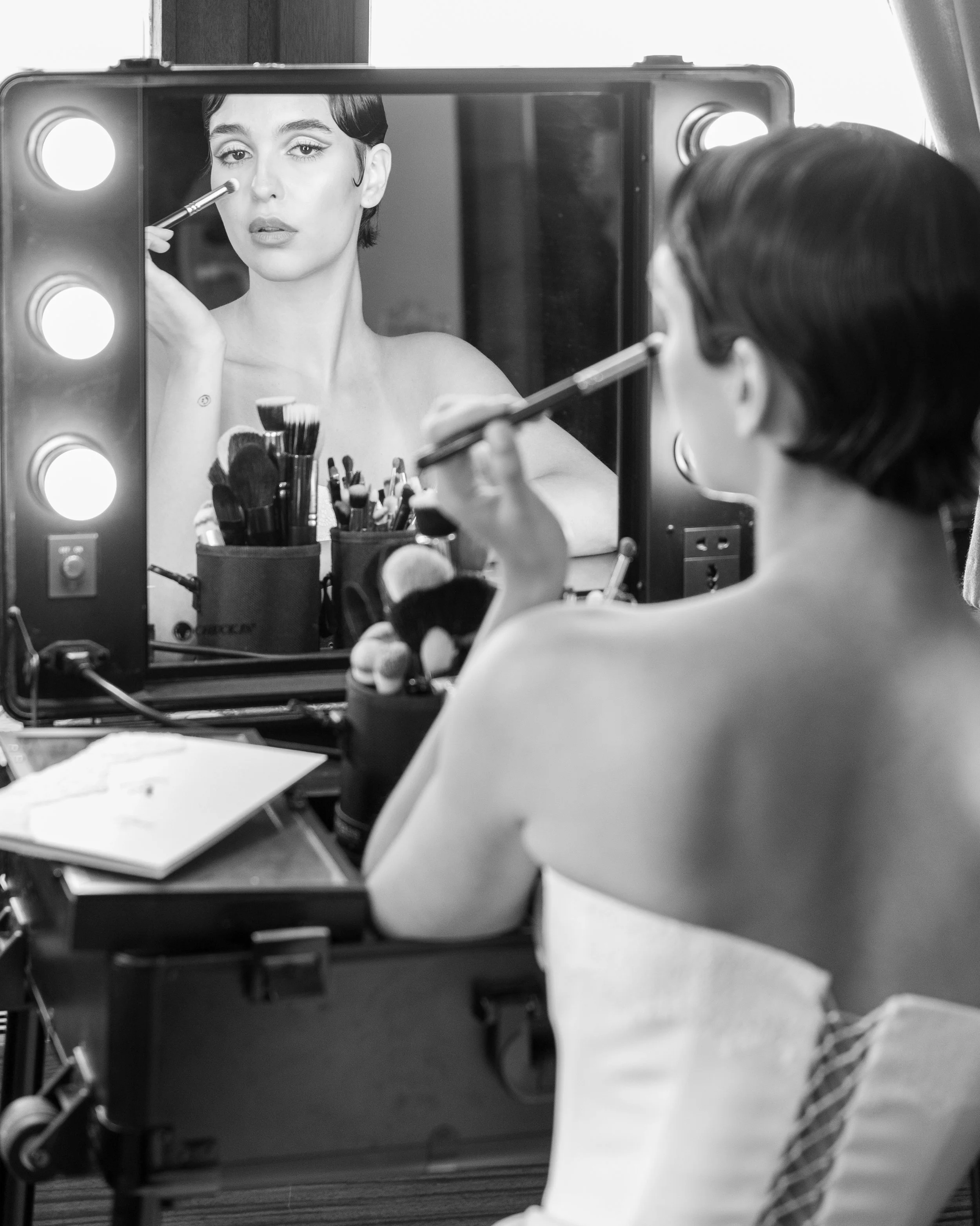 A person with dark hair and light skin applying makeup to a woman’s face in front of a mirror with lighting bulbs. The makeup tools are arranged in containers on the table.