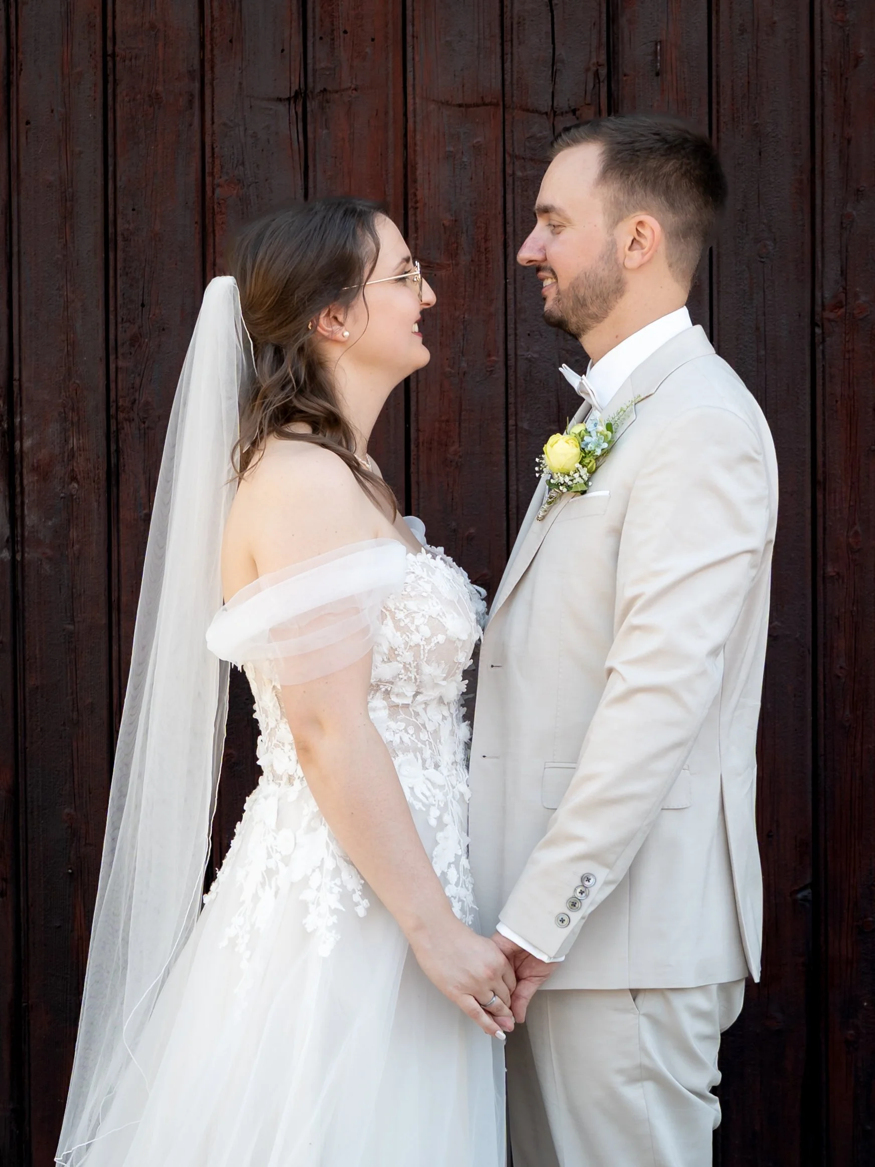 A bride and groom dressed in wedding attire are standing face to face, holding hands, and looking at each other with smiles against a wooden background.
