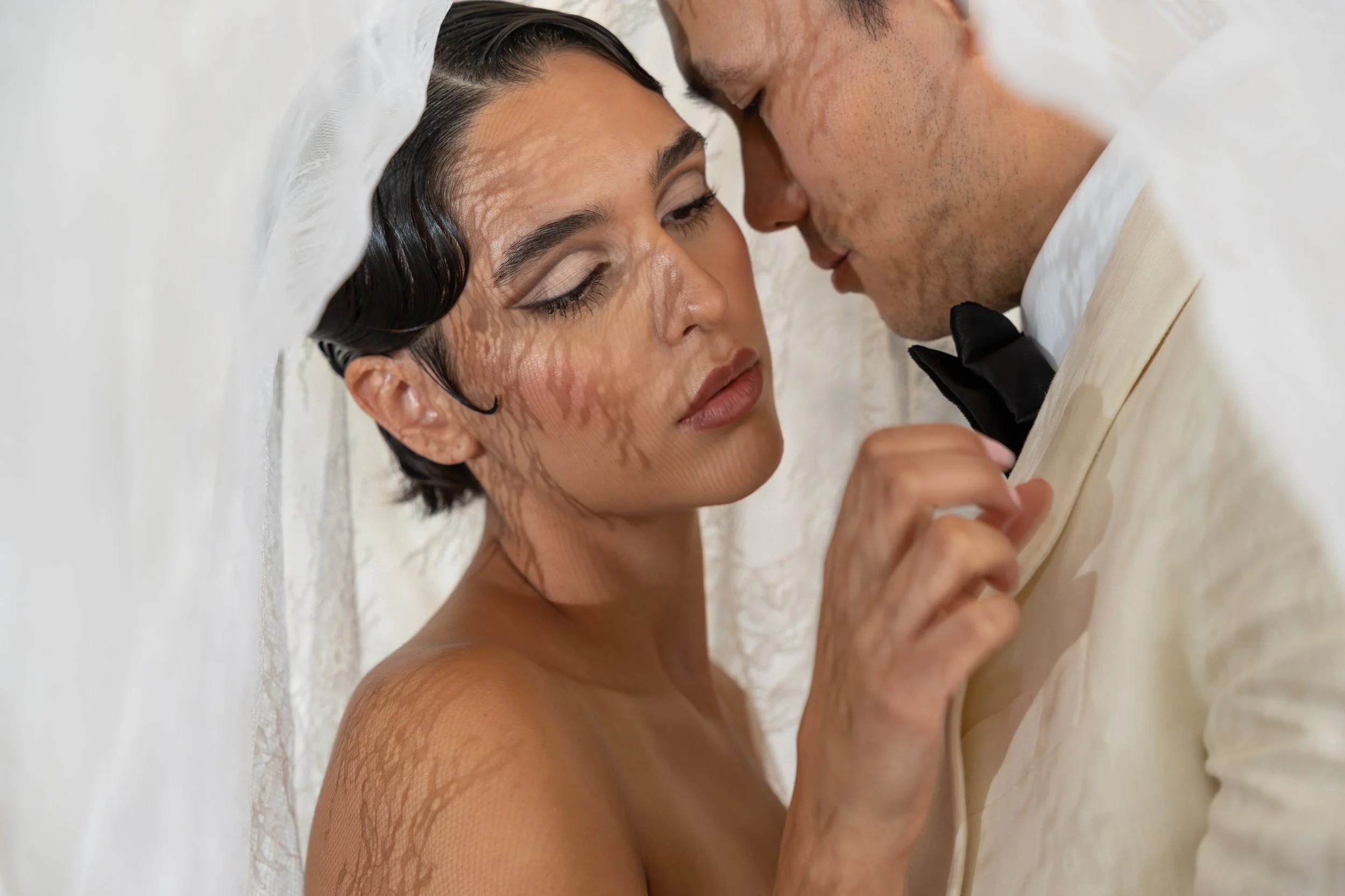 A couple faces close to each other, with the woman lying on a white surface and the man leaning over her, both dressed in formal attire, with the woman wearing makeup and the man in a tuxedo.