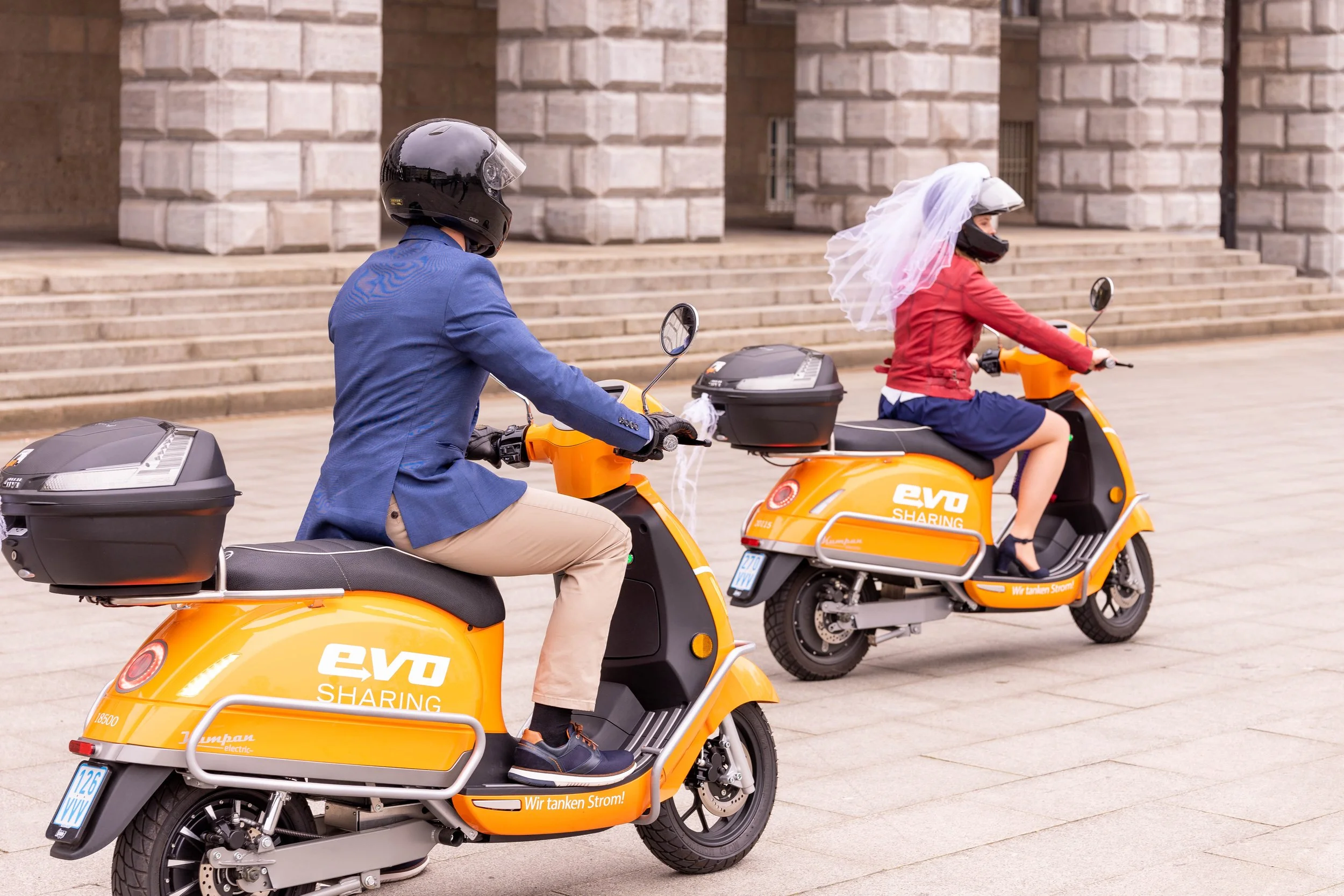 Two people riding orange electric scooters in front of a stone building, with one person wearing a bridal veil and dresses, indicating a possible themed ride or celebration.