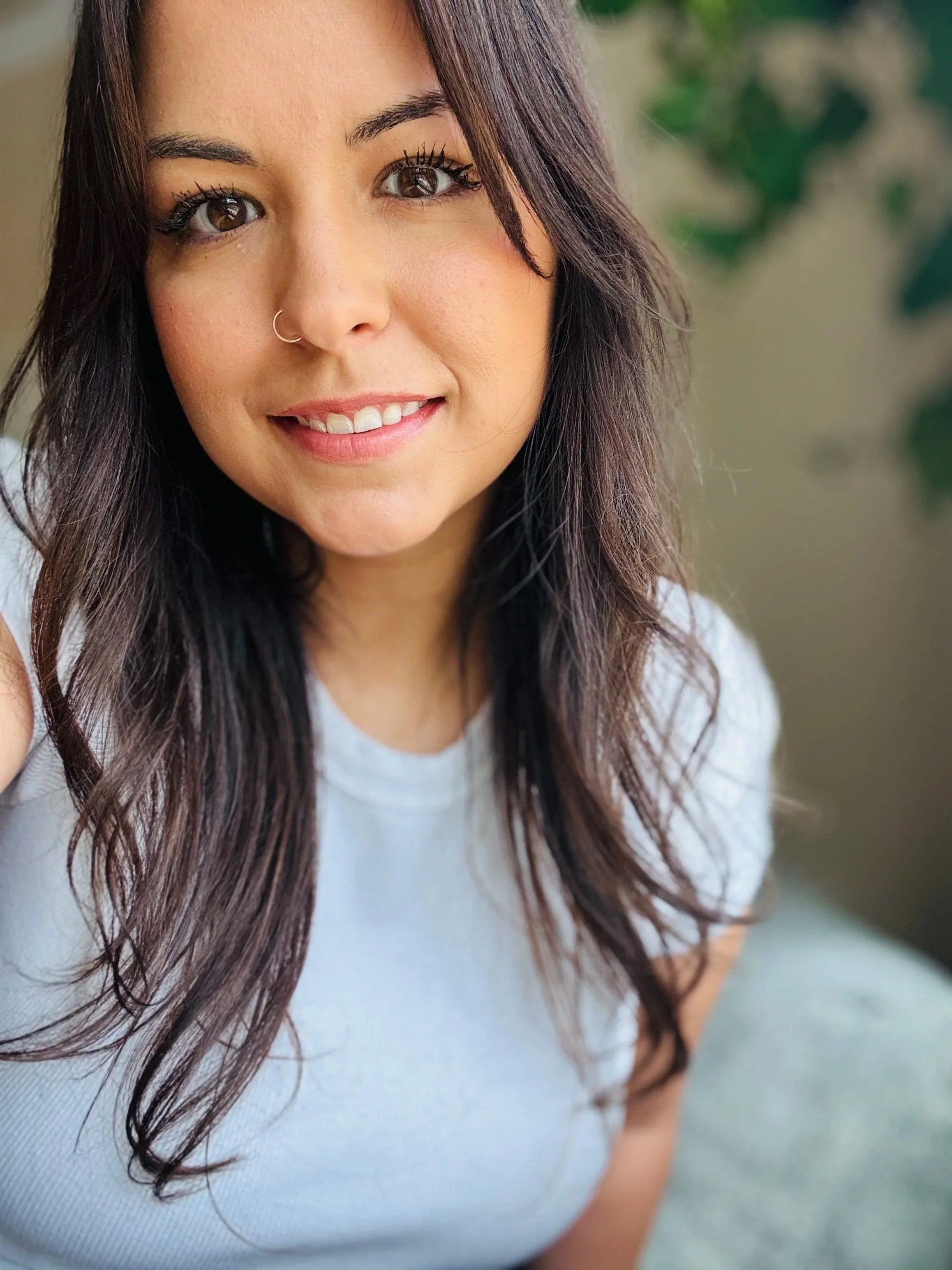 Close-up of a young woman with wavy dark hair and brown eyes, smiling and wearing a white top.