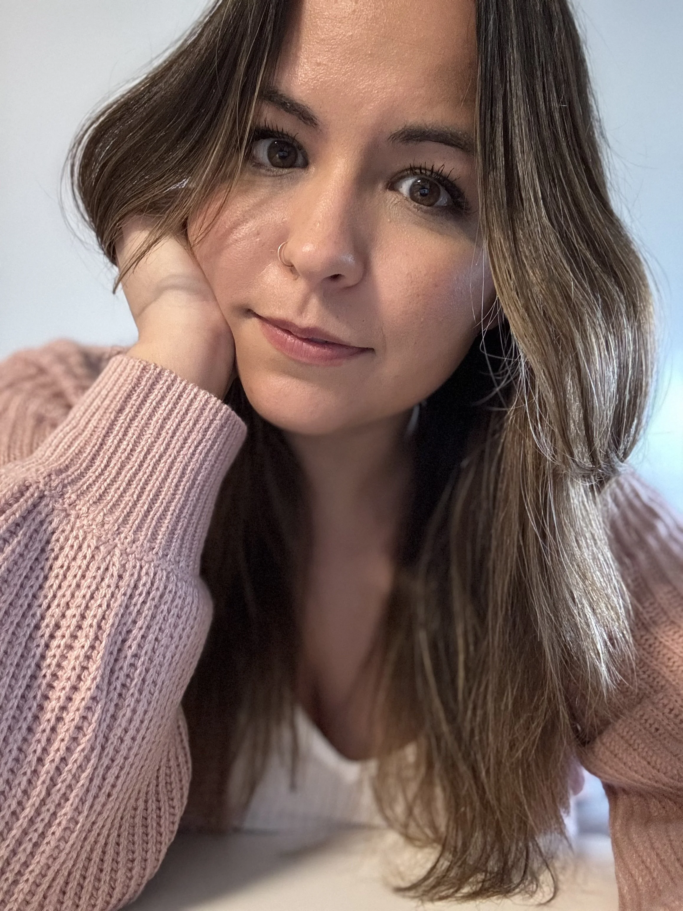 Close-up of a woman with long brown hair, brown eyes, and a nose ring resting her face on her hand while wearing a pink sweater.