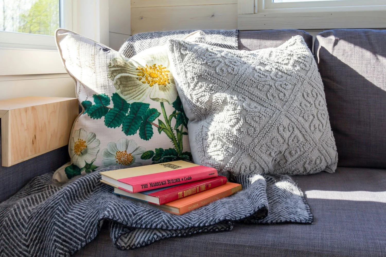 A cozy living room corner with a gray couch, decorated with a floral pillow, a textured gray pillow, and three books stacked on a gray blanket. There is a wooden side table next to the couch and a large window letting in natural light.