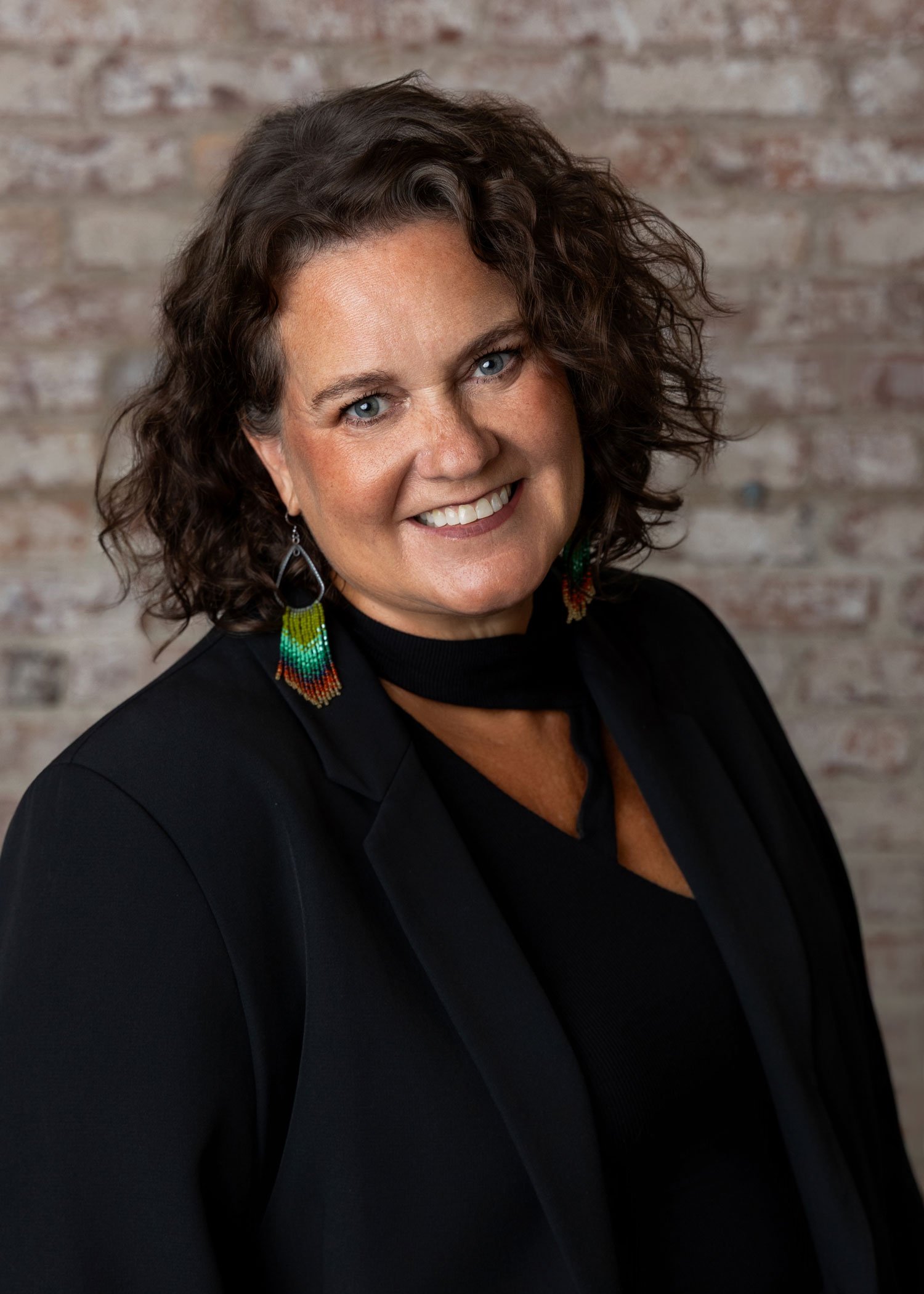 Jennifer Noteboom, a woman with curly brown hair, blue eyes, smiling, wearing colorful beaded earrings and a black top, standing against a brick wall background.