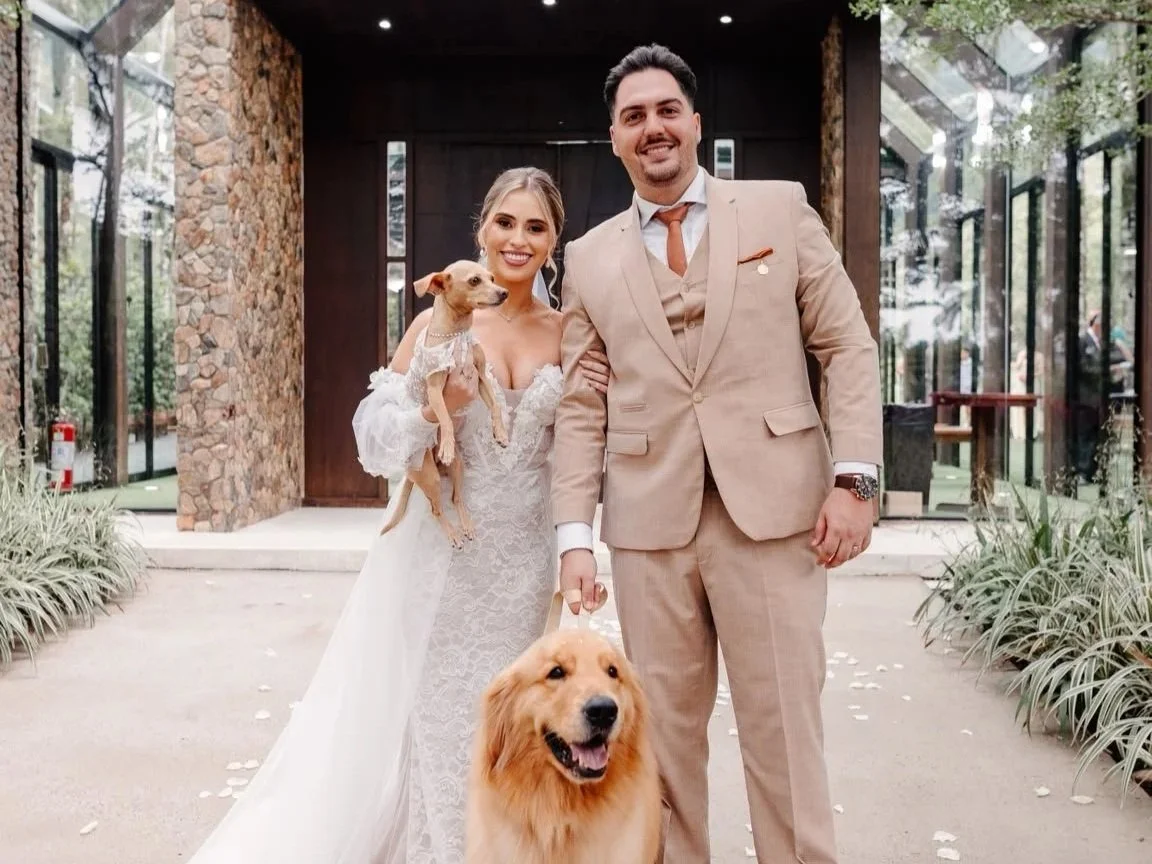 Casal sorridente em casamento ao ar livre com dois cães, um pequeno na mão da mulher e um grande no chão.