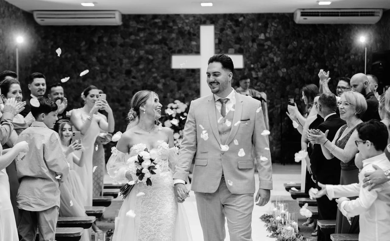 Casal de noivos caminhando de mãos dadas pelo corredor ao lado de convidados em uma cerimônia de casamento, decorada com flores e uma cruz ao fundo.