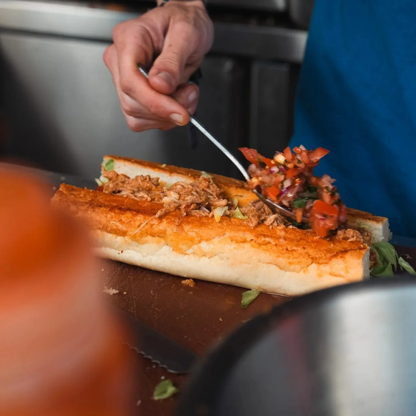 A person is scooping shredded meat and diced vegetables onto a sandwich with a spoon. The sandwich has a long white bun and ingredients inside, on a wooden cutting board.