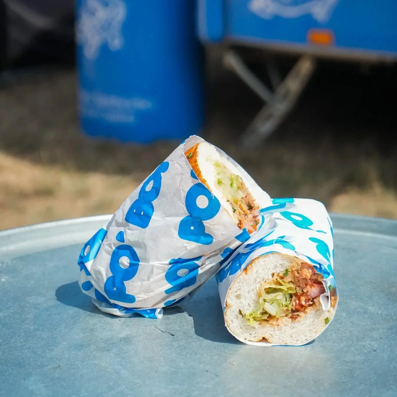 A wrap sandwich with lettuce, tomato, and meat in crumpled blue and white paper sitting on a gray surface, with a blurred blue object and truck in the background.
