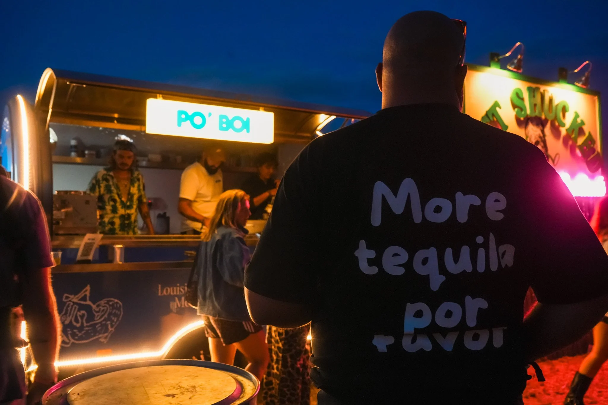 Night scene at a fair or carnival with a person wearing a black shirt with partially visible text, people in the background at a food stand, and colorful lights and signs.