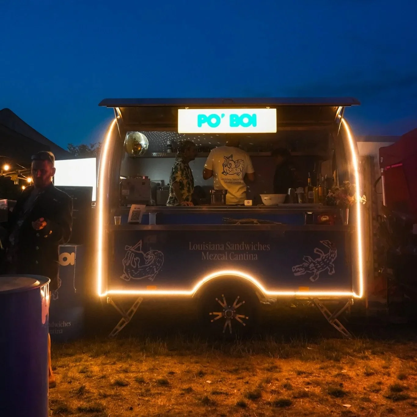A food truck at night serving Louisiana sandwiches with a sign above and oval lights outlining the truck's edges. People are inside preparing food, and a man stands outside on the left.