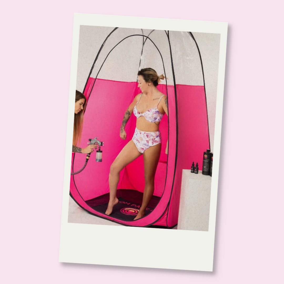 Woman in floral swimsuit standing inside a pink spray tan tent while a woman sprays her with a spray tan.