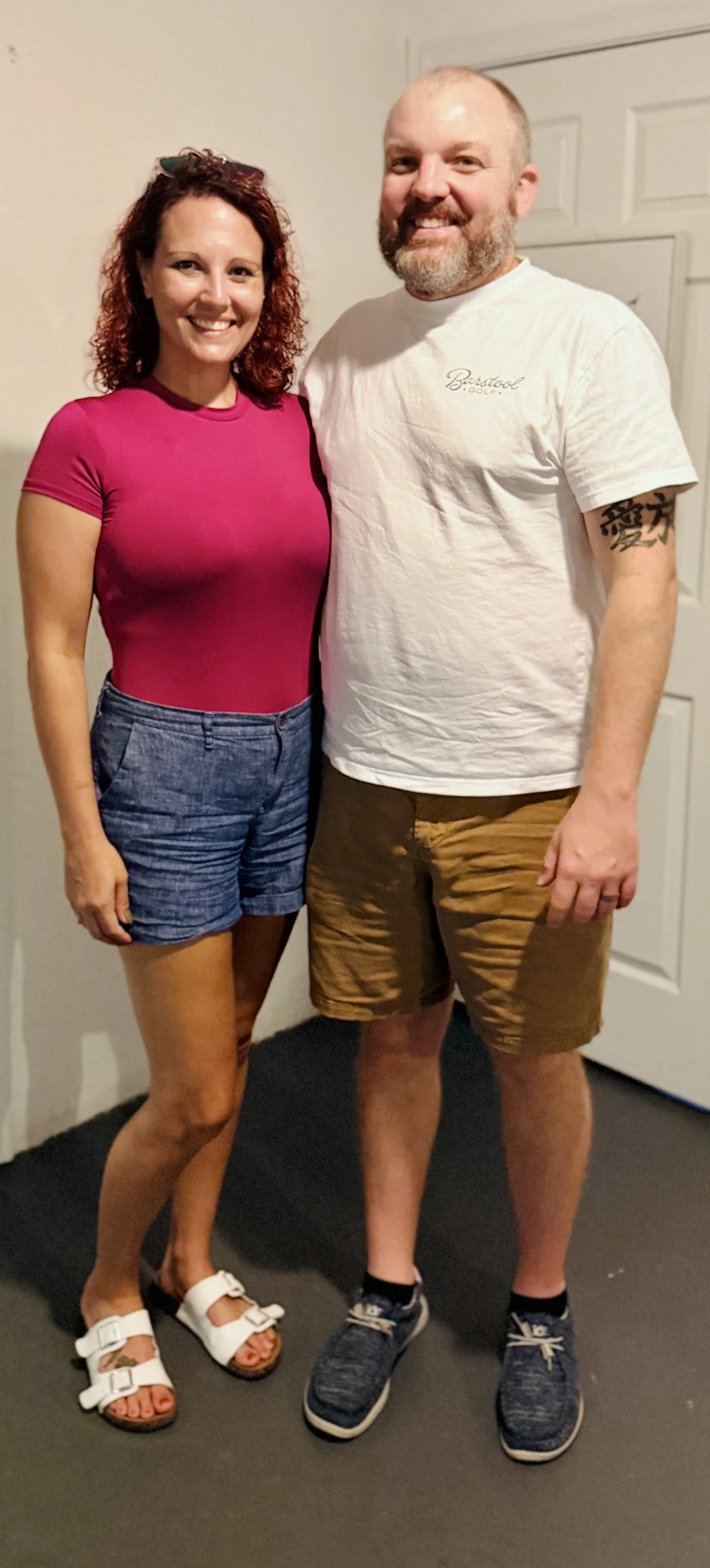 A smiling woman with curly red hair wearing a pink shirt and denim shorts, standing next to a smiling man with a beard wearing a white T-shirt and brown shorts, inside a house near a white door.