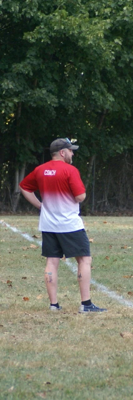 A man wearing a 'COACH' shirt, shorts, a cap, and sunglasses standing on a grassy field near trees.