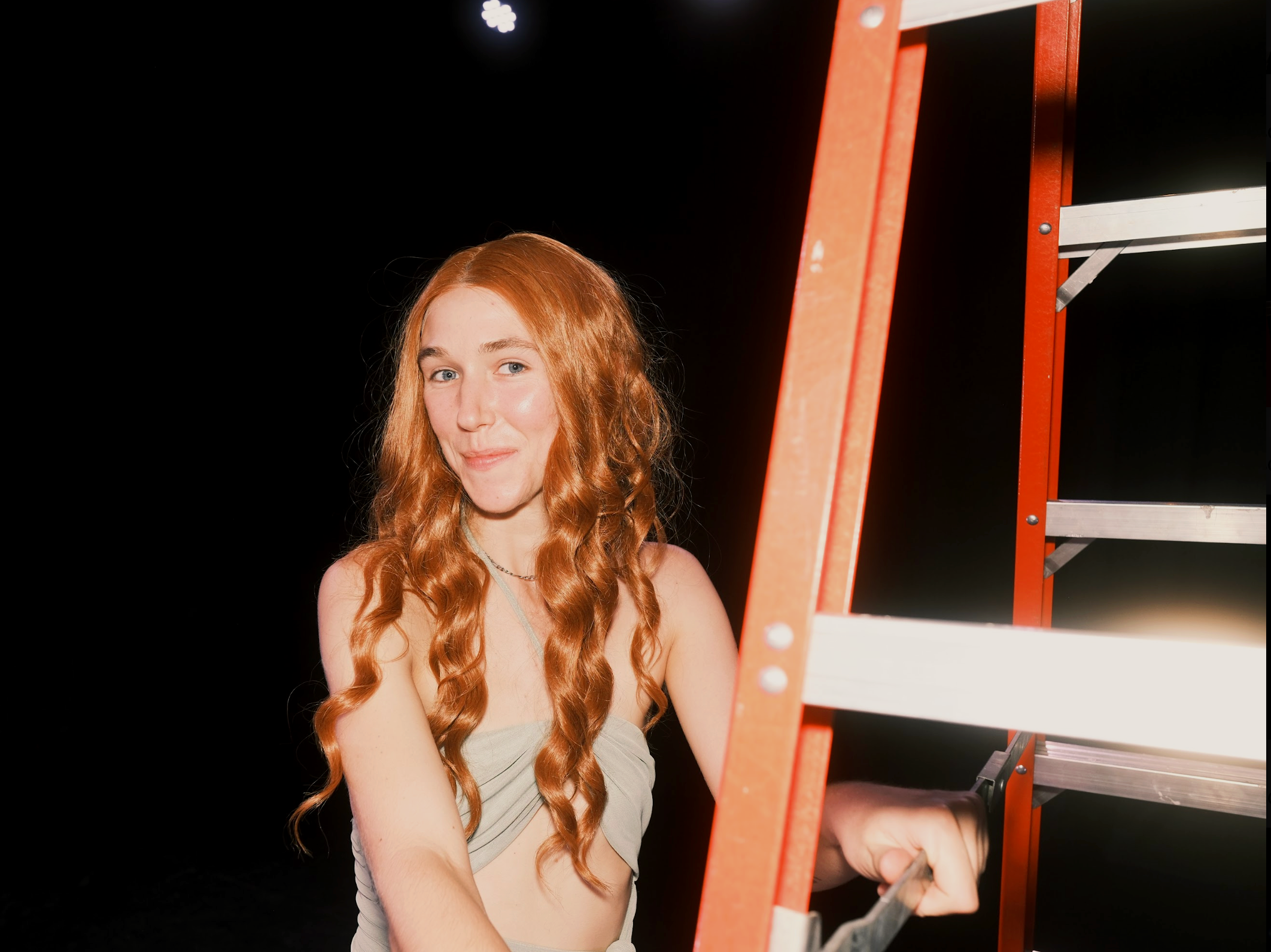 A young woman with long red hair and a sage-colored performance dress standing next to a red ladder in a dark theater.
