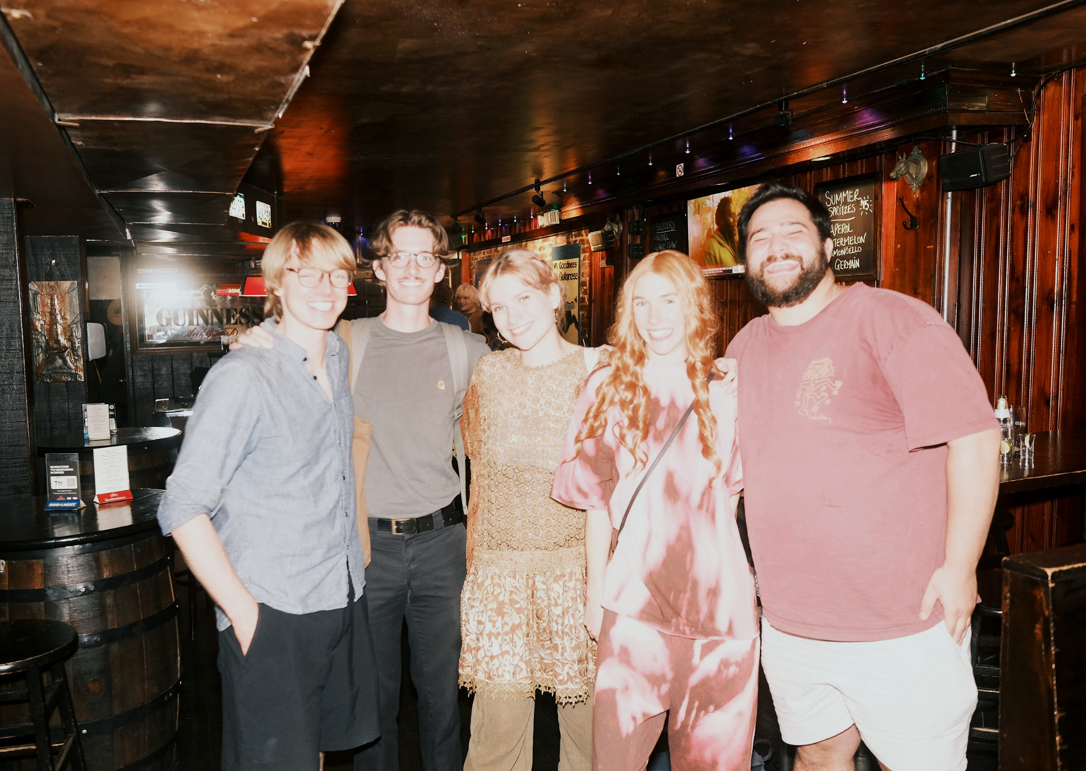 Group of five young adults smiling and posing together inside a pub with dark wooden decor.